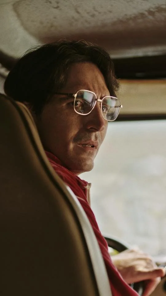 A man with glasses, dark hair, and a red shirt sits in a vehicle, looking over his shoulder towards the camera with a serious expression.