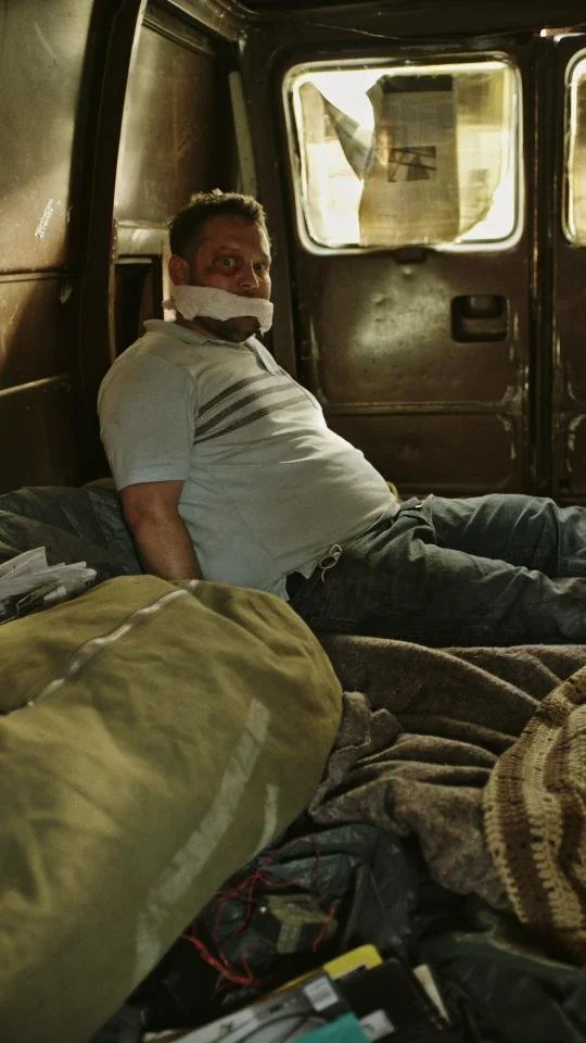 Man sitting inside a dark, enclosed space with windows, having a cloth gag over his mouth, wearing a light-colored shirt and dark pants.