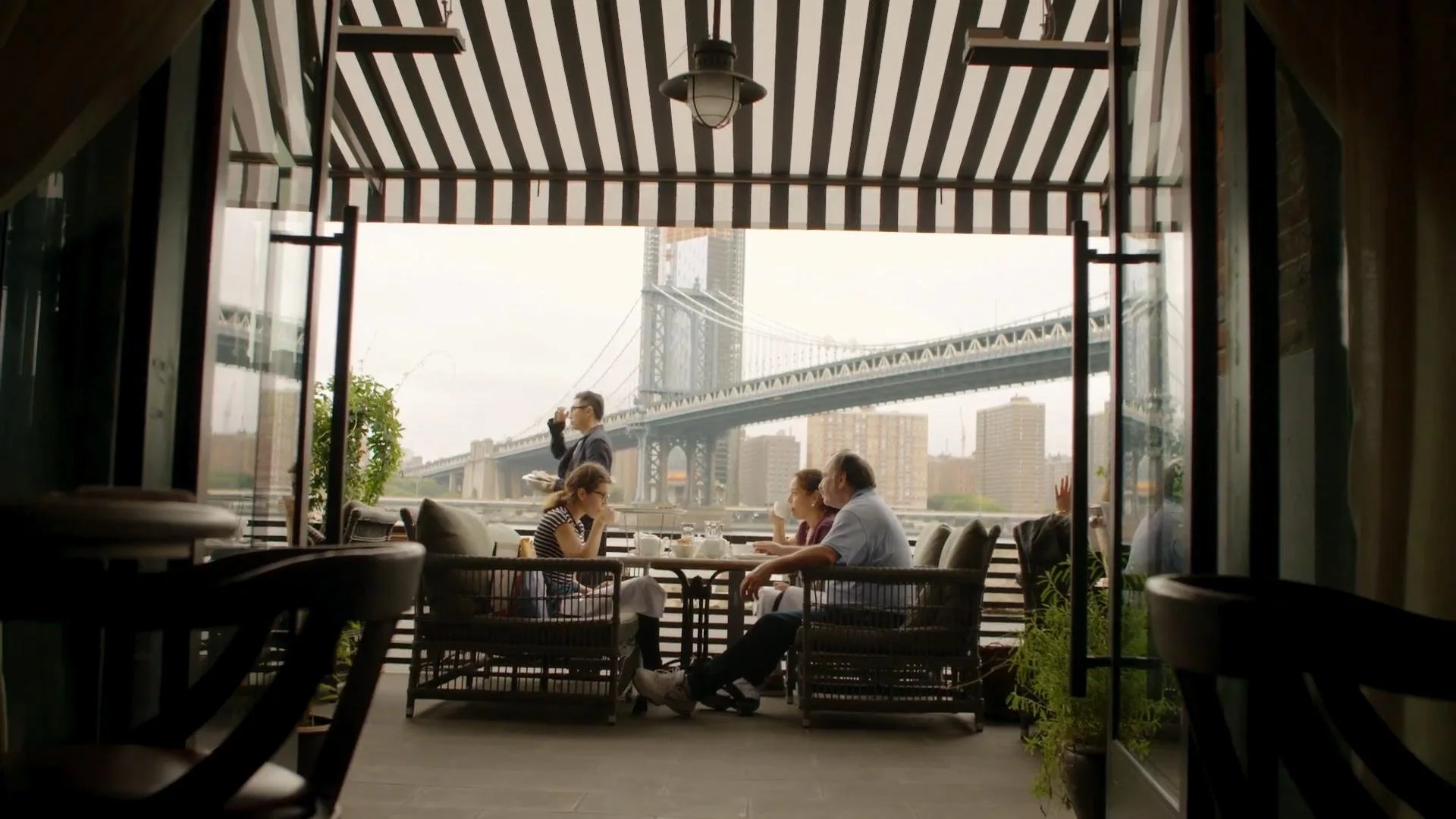 A view from inside a restaurant with an open patio, overlooking the Brooklyn Bridge in New York City. Four people are seated at a table, engaging in conversation. A waiter stands nearby, holding a menu. The scene is framed by black-and-white striped 