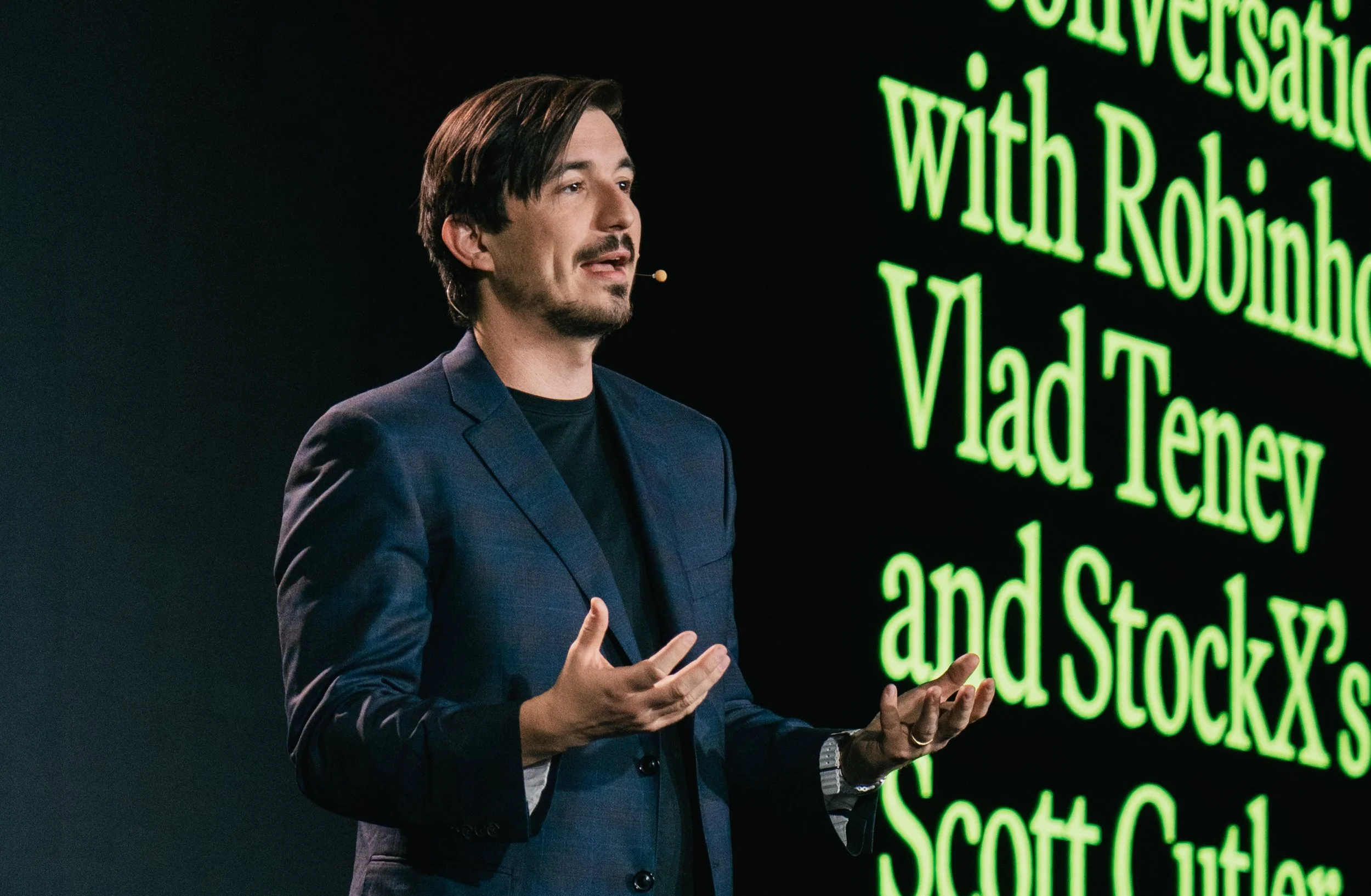 A man with dark hair and a beard dressed in a navy blazer and black shirt giving a presentation with a green text display behind him.