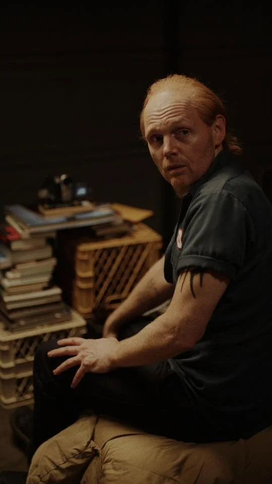 A man with reddish hair and tattoos on his arm, sitting on a beige cushion in a dimly lit room, with a stack of books and papers on a wooden table in the background.