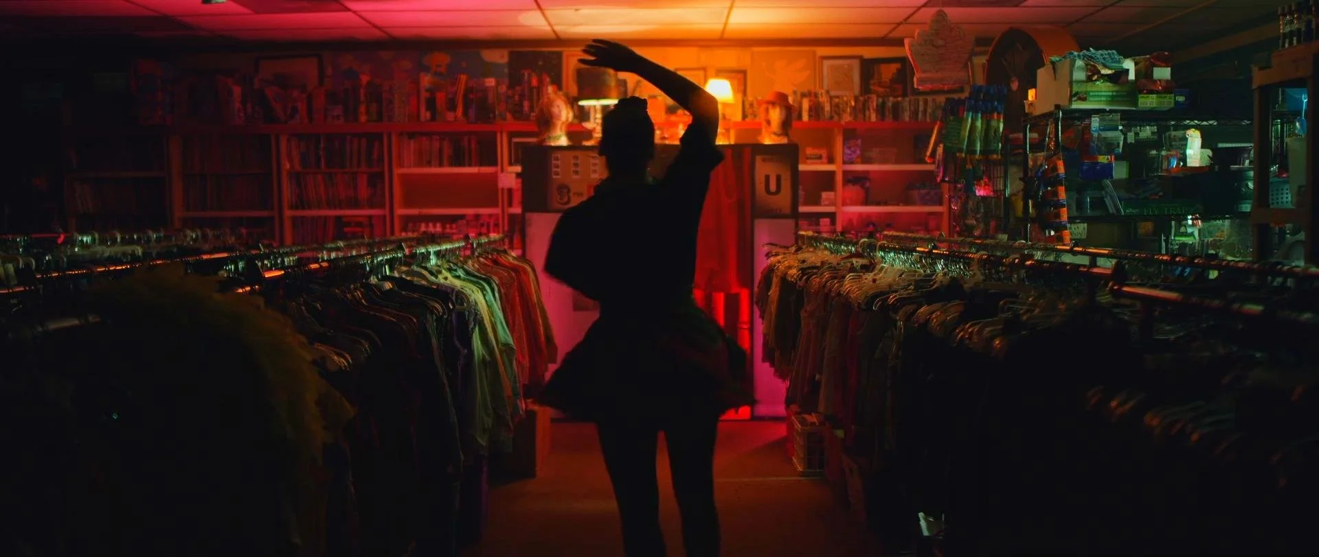 A person inside a dimly lit thrift store browsing through clothes on racks, illuminated by a warm orange light.