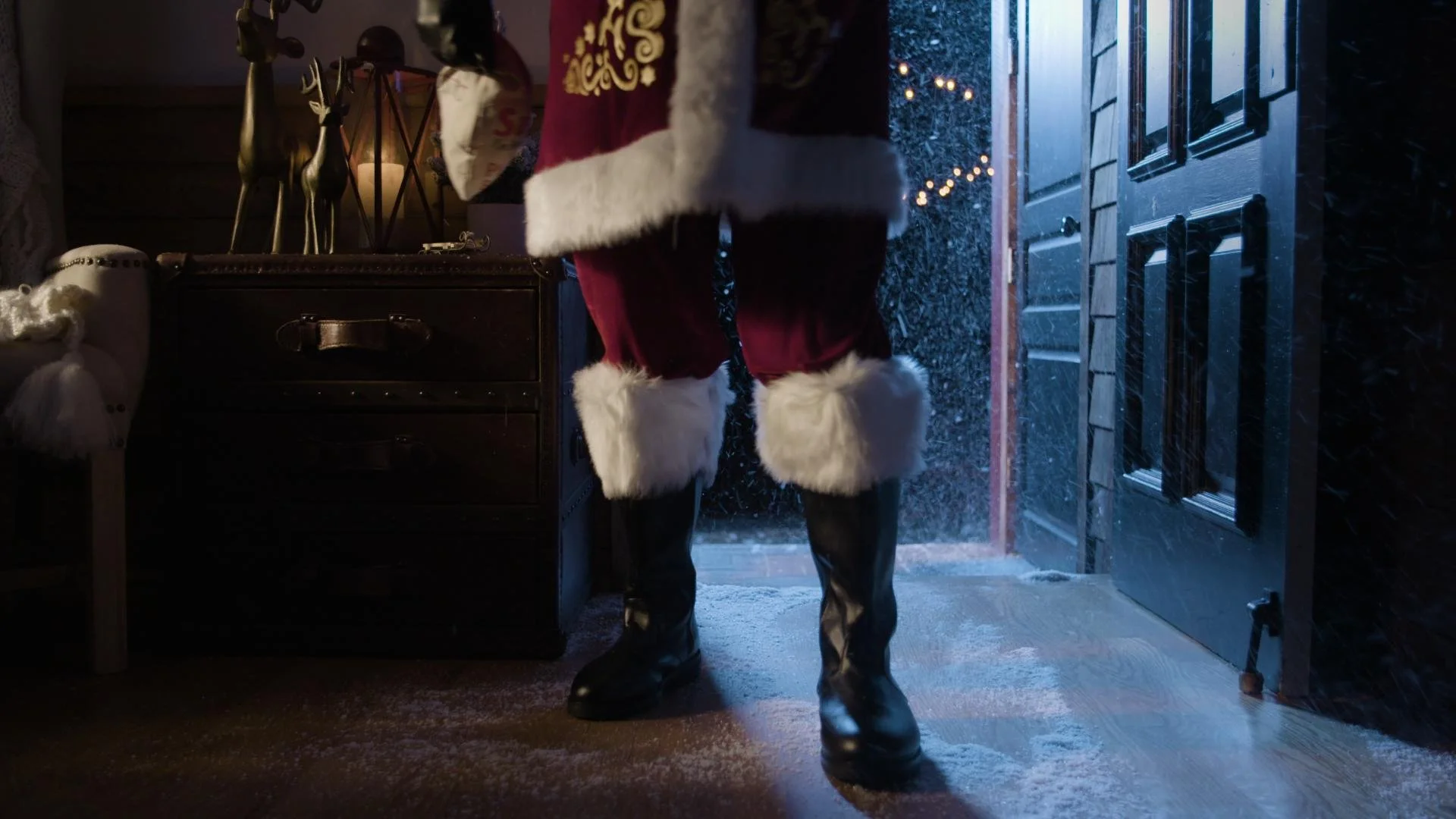Person dressed as Santa Claus standing inside a house near the open front door on a snowy night.