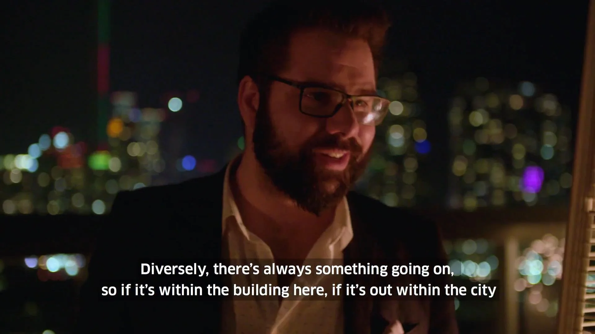 A man with glasses and a beard smiling at night, with a city skyline illuminated in the background and subtitles reading, 'Diverse, there's always something going on, so if it's within the building here, if it's out within the city.'