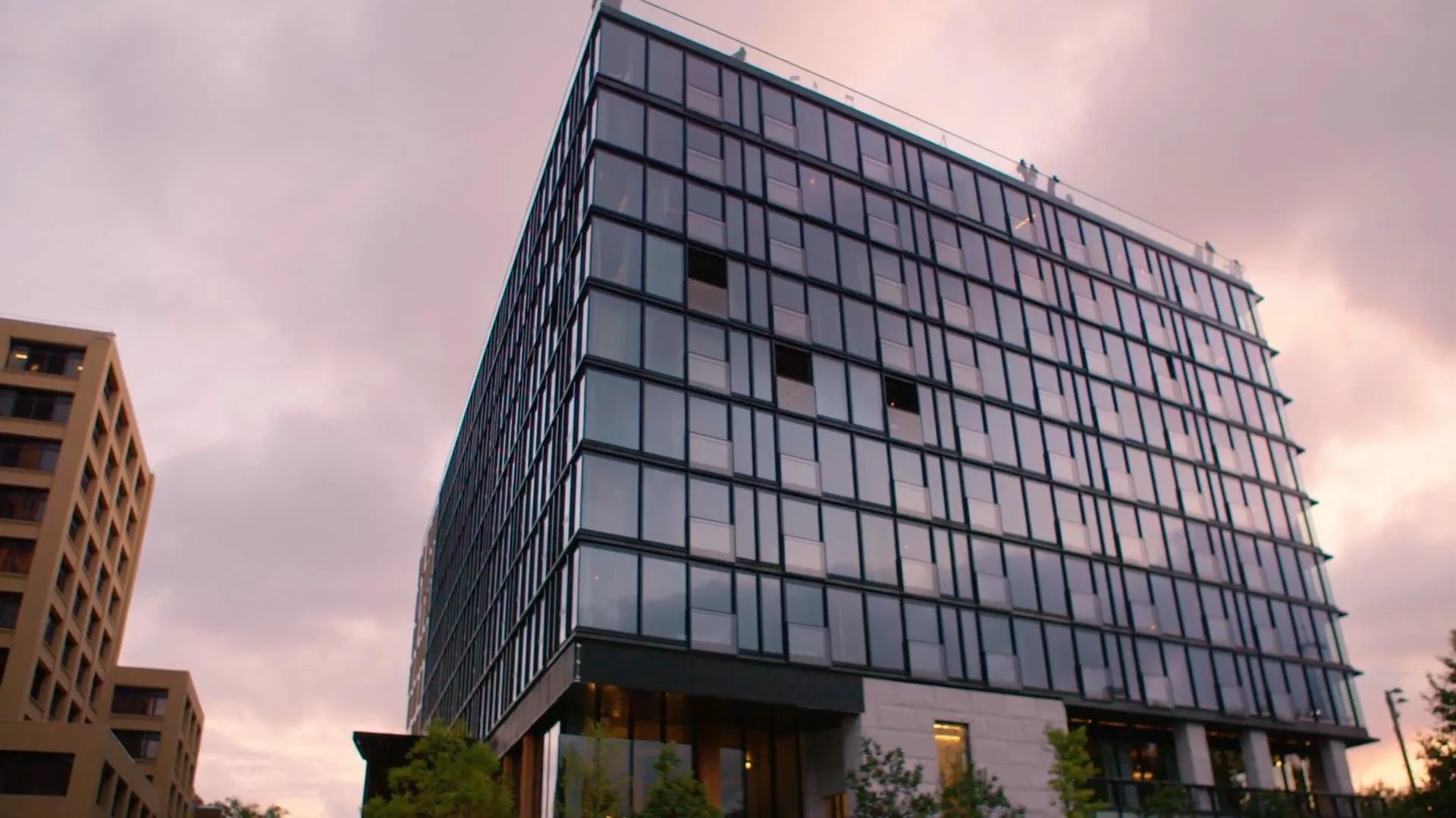 A modern multi-story glass building with a grid facade, situated in an urban area during dusk with a pinkish sky, and some trees at the bottom.