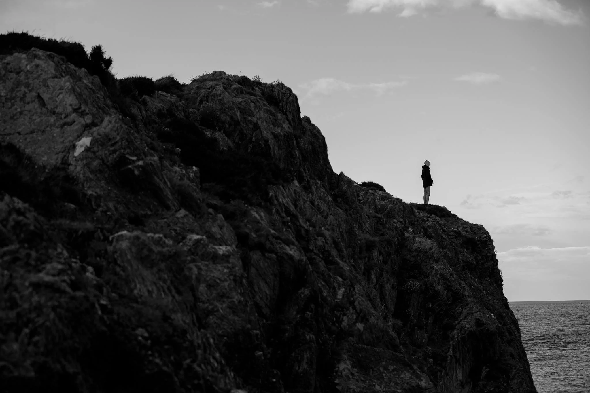 A black-and-white photo of a person standing on a rocky cliff overlooking the ocean with cloudy sky in the background.