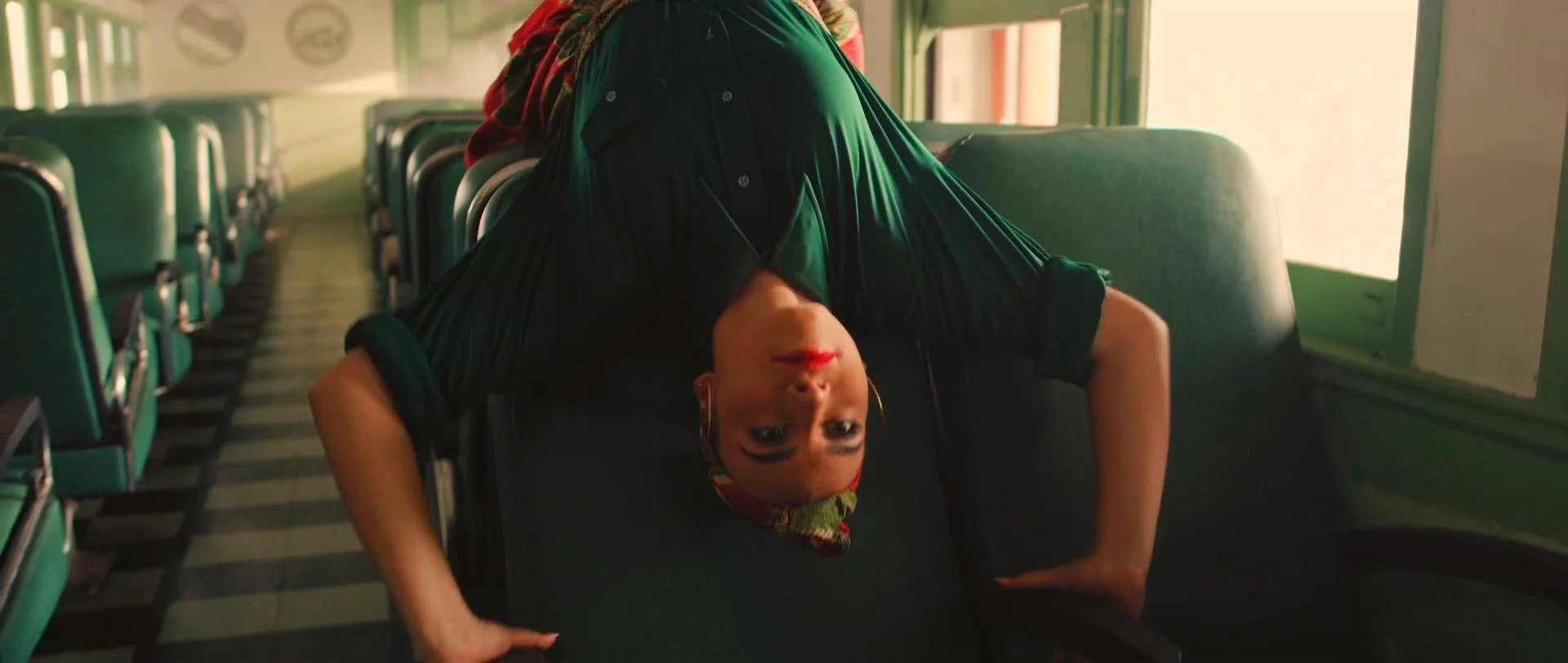 A woman with dark hair and red lipstick is upside down on a chair in a vintage bus or train, with rows of green seats and sunlight coming through the window.