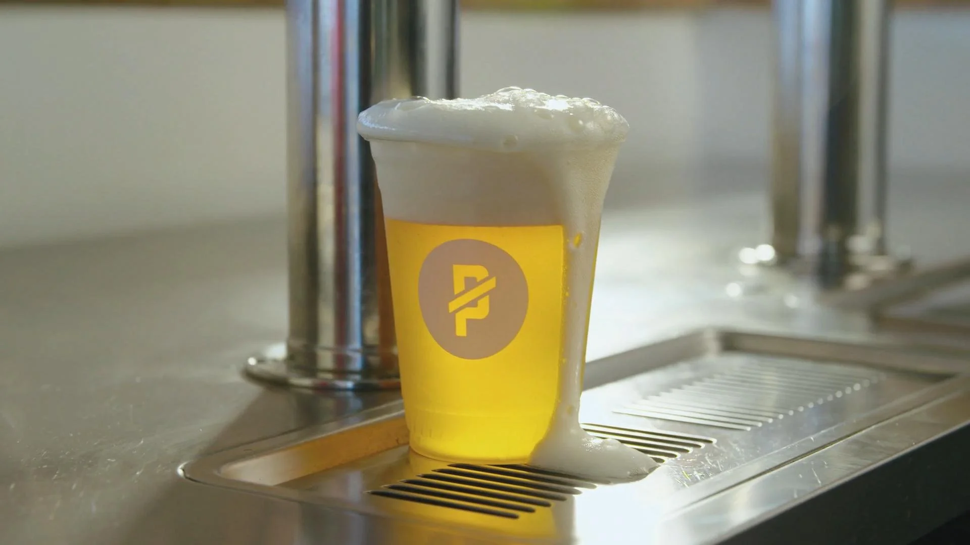 A yellow paper coffee cup with white foam overflowing and dripping down the sides, placed under a coffee machine spout in a cafe setting.
