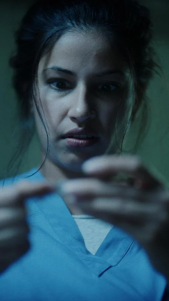 A woman with dark hair pulled back, wearing blue medical scrubs, looking intently at a small object she is holding with both hands.