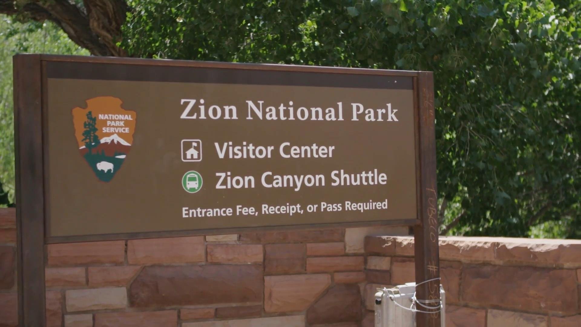 Sign for Zion National Park displaying the National Park Service logo, and information about a Visitor Center and Zion Canyon Shuttle, with a note about entrance fee, receipt, or pass requirements.