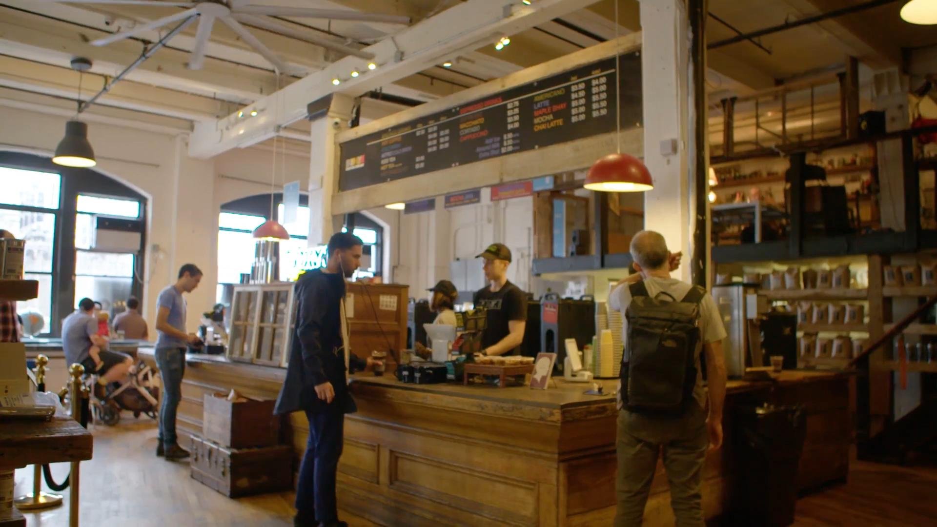 Interior of a coffee shop with a wooden counter, baristas serving customers, several people sitting and standing, large windows, and industrial-style lighting.
