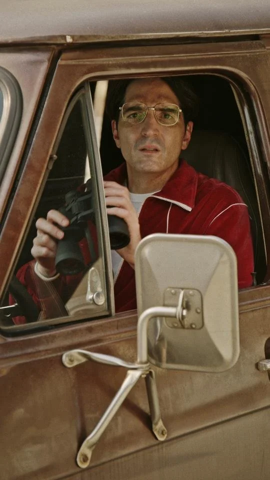 Man with glasses sitting in a vintage vehicle, holding binoculars, wearing a red jacket and a white shirt.