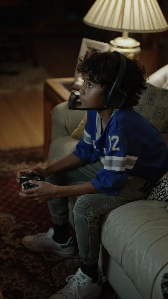 Boy with curly hair wearing a blue sports jersey, sitting on a beige couch, holding a game controller, with a gaming headset around his neck, in a cozy living room with a lamp and pictures on the table behind him.
