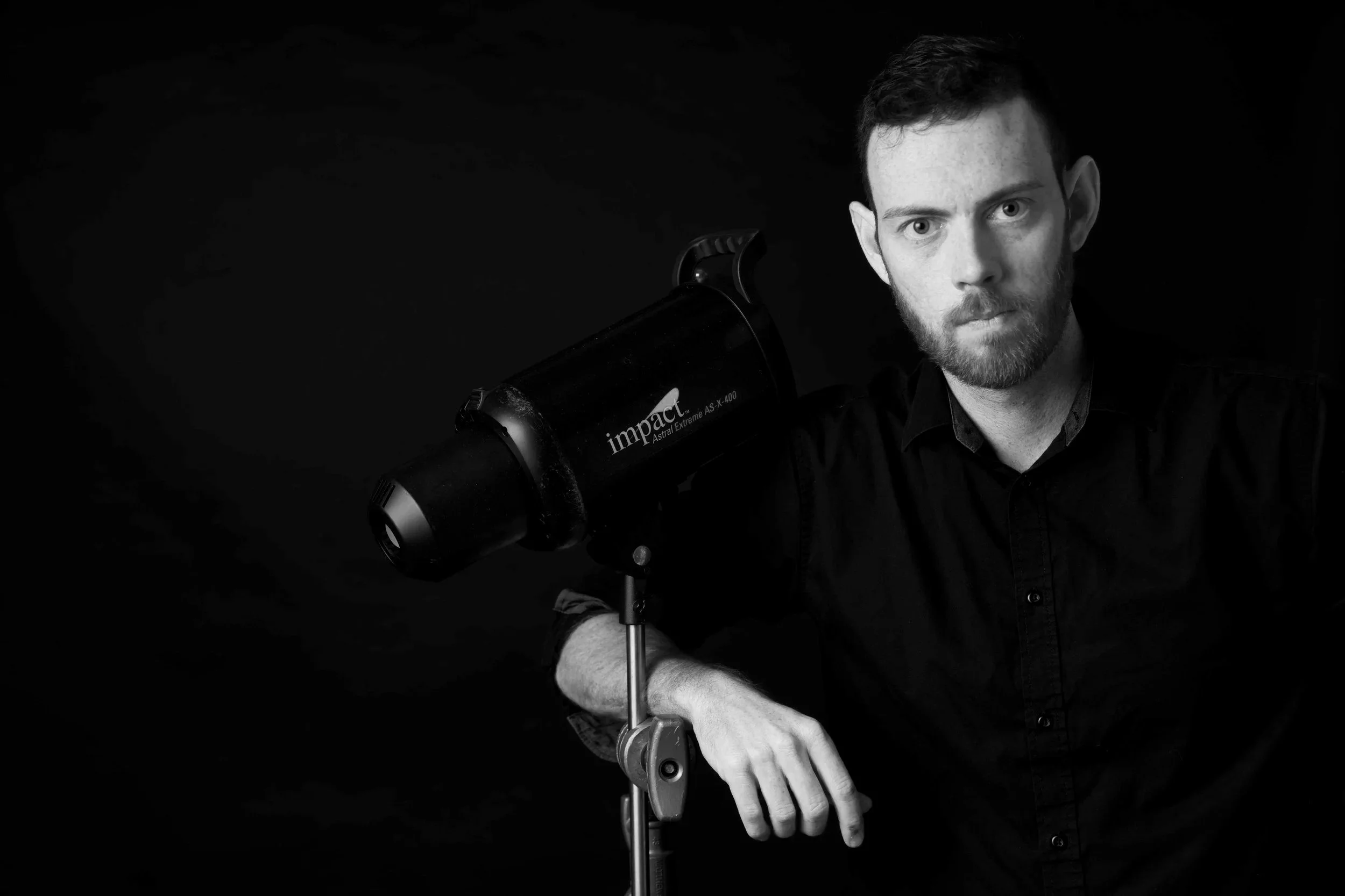 A black and white photo of a man with a beard and short hair, wearing a dark shirt. He is looking at the camera with a serious expression and is positioned next to a telescope labeled "Impact Astral Extreme AS-X-400". He is standing against a dark background.