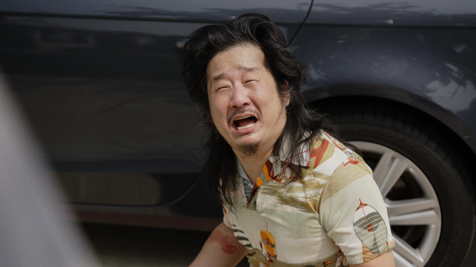 A man with long black hair and a goatee, wearing a colorful vintage shirt, appears to be in distress or pain and is on the ground near a black car with a tire visible in the background.