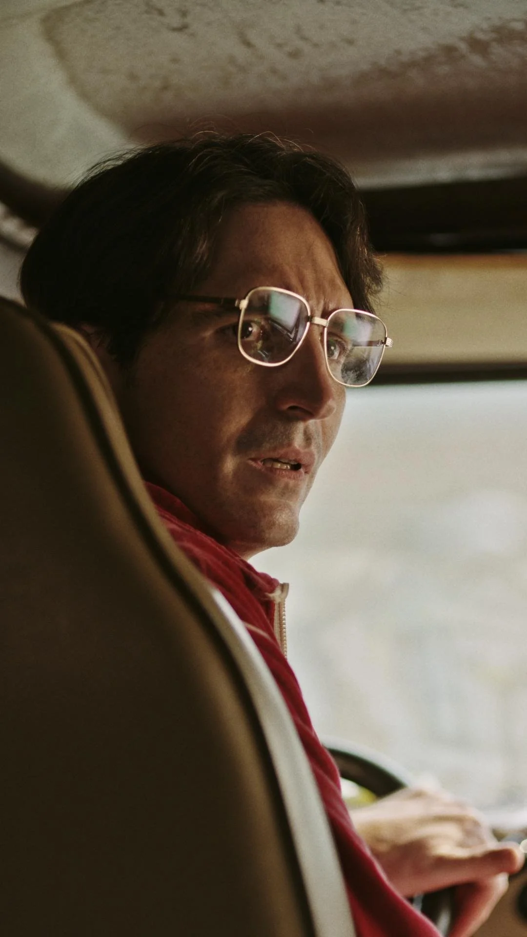 A man with glasses wearing a red jacket is sitting on a bus or car seat, looking towards the window.