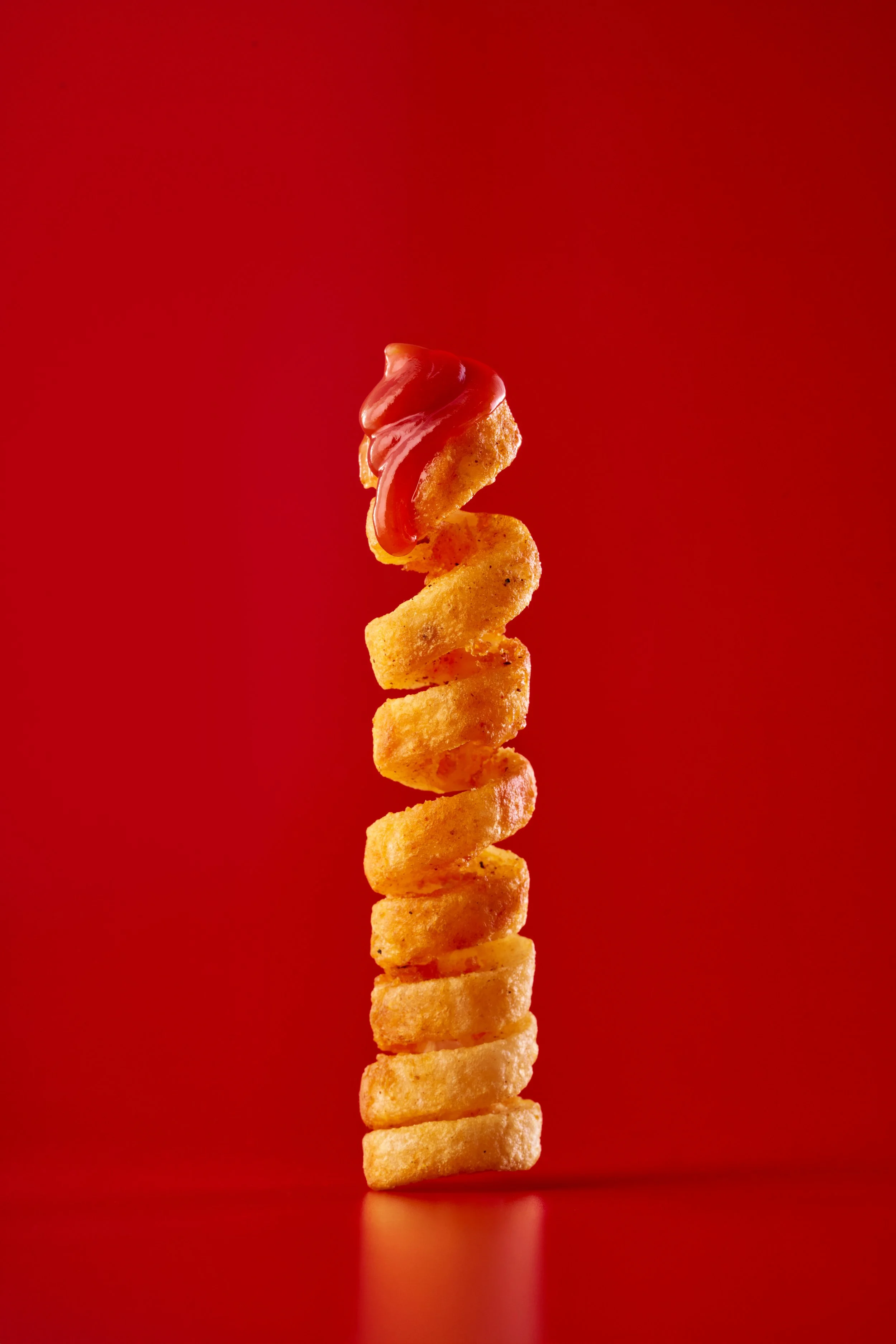 A vertical stack of curly fries topped with ketchup on a red background.