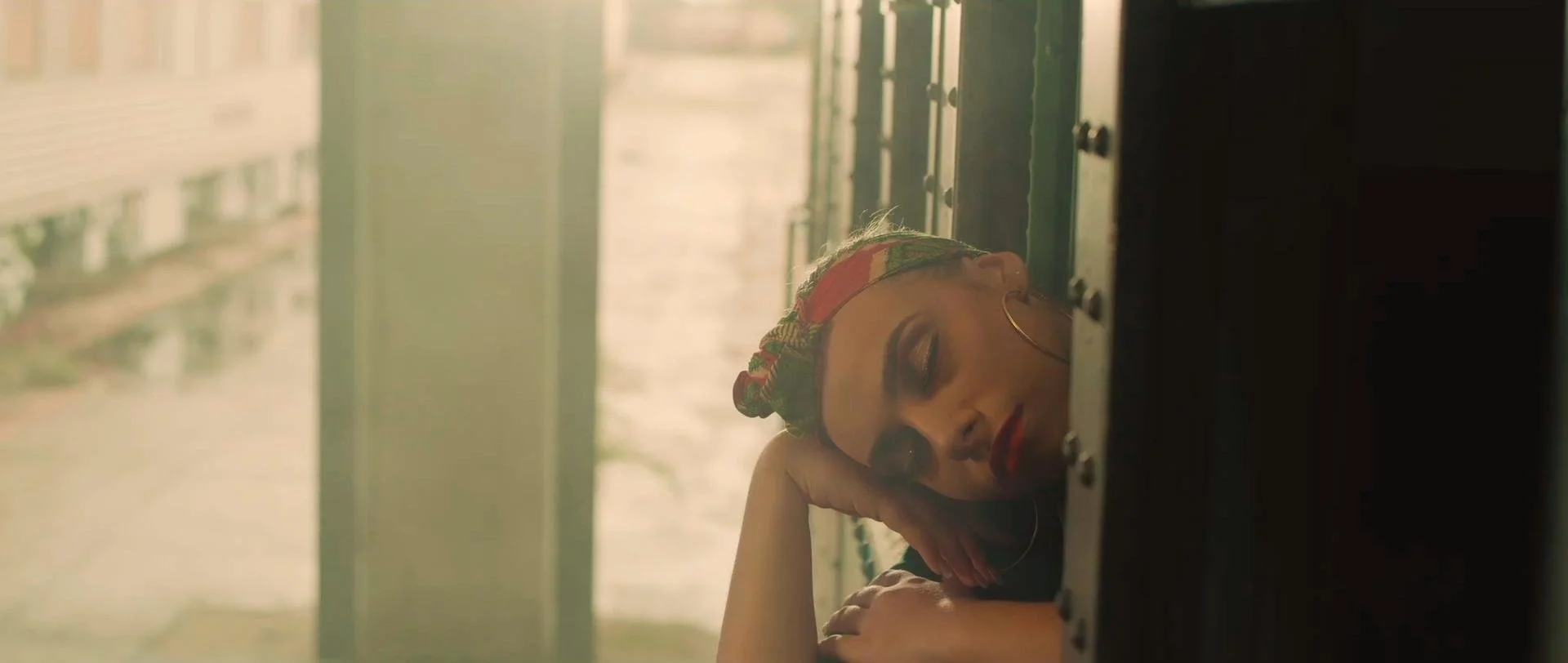 A woman with a colorful headscarf and hoop earrings is resting her head and hand on a table, eyes closed, in a room with a blurred background.