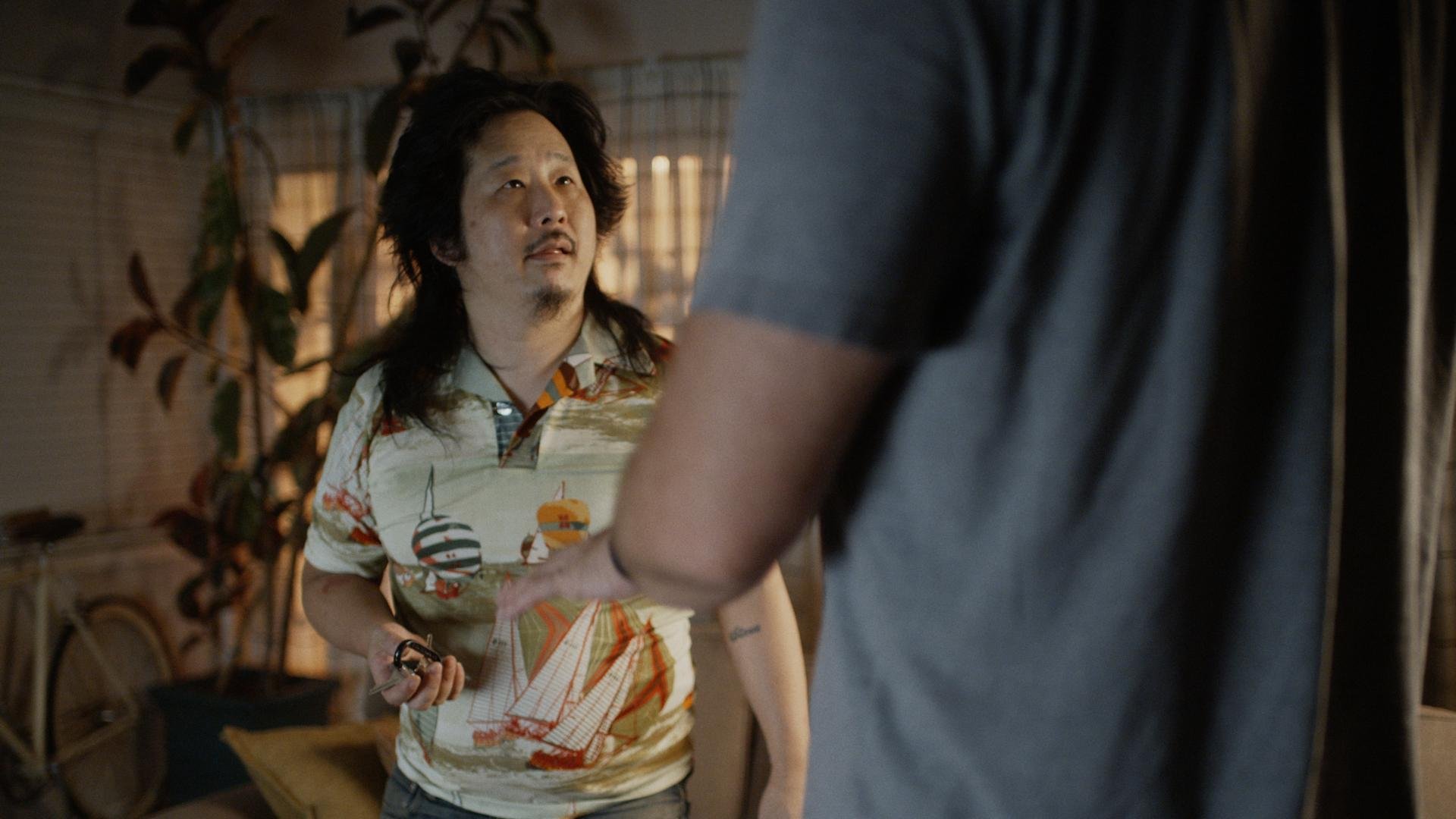 A man with long dark hair and a floral shirt is seen talking to someone not fully visible in a dimly lit indoor space.