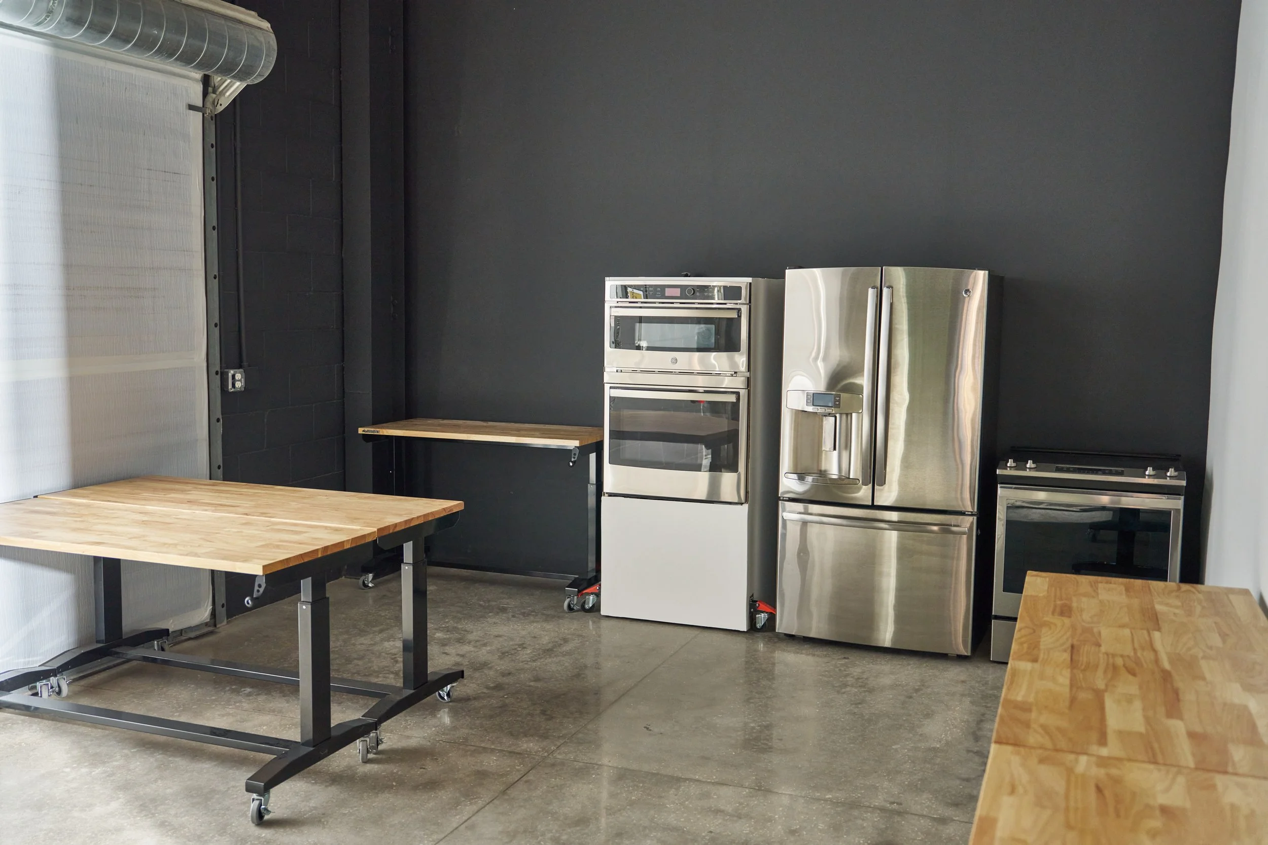 Stainless steel refrigerator and oven in a modern kitchen with black and white walls, wooden tables, and concrete floors.