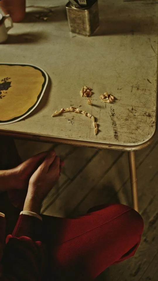 A wooden table with a few small crust remnants, a partially peeled banana, and a metal container. A person's hand resting near the table, wearing a dark red sleeve.