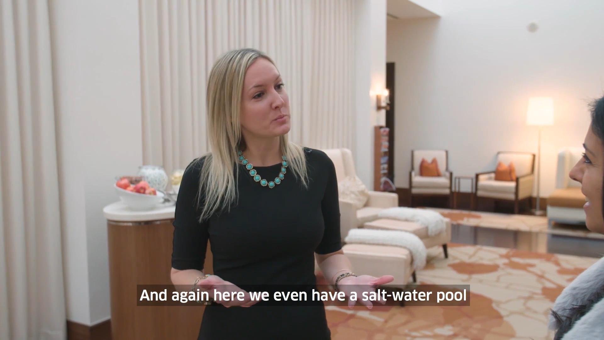 A woman with blonde hair wearing a black dress and turquoise necklace talking to a man in a brightly lit room with cream-colored walls and furniture, with a subtitle that reads 'And again here we even have a salt-water pool'.