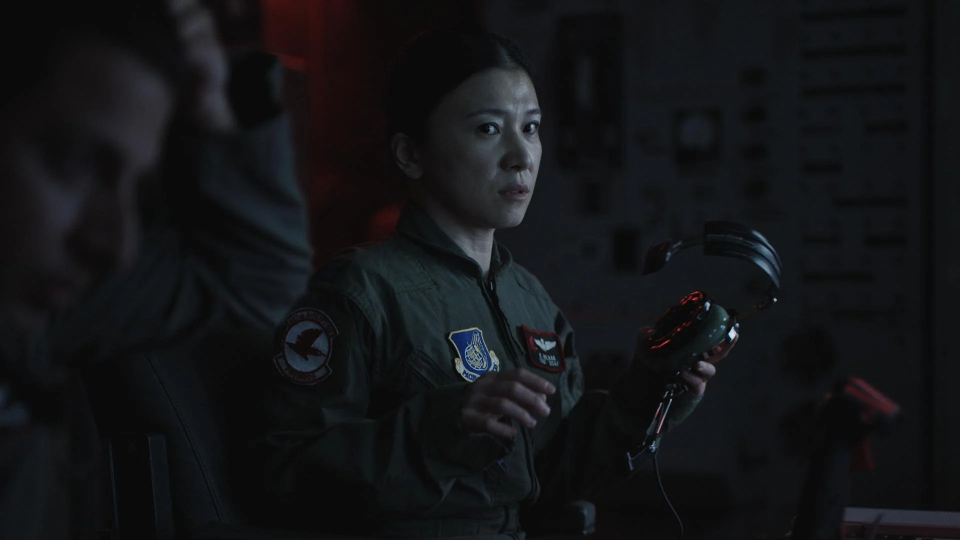 A woman in military uniform holding a headset in a dark room with a serious expression.