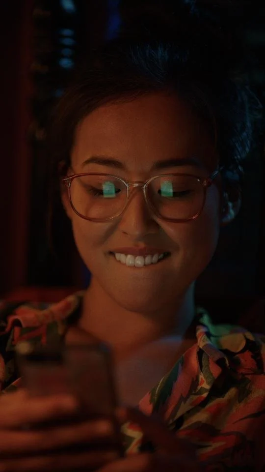 Young woman with glasses smiling while looking at her phone in a dimly lit room.