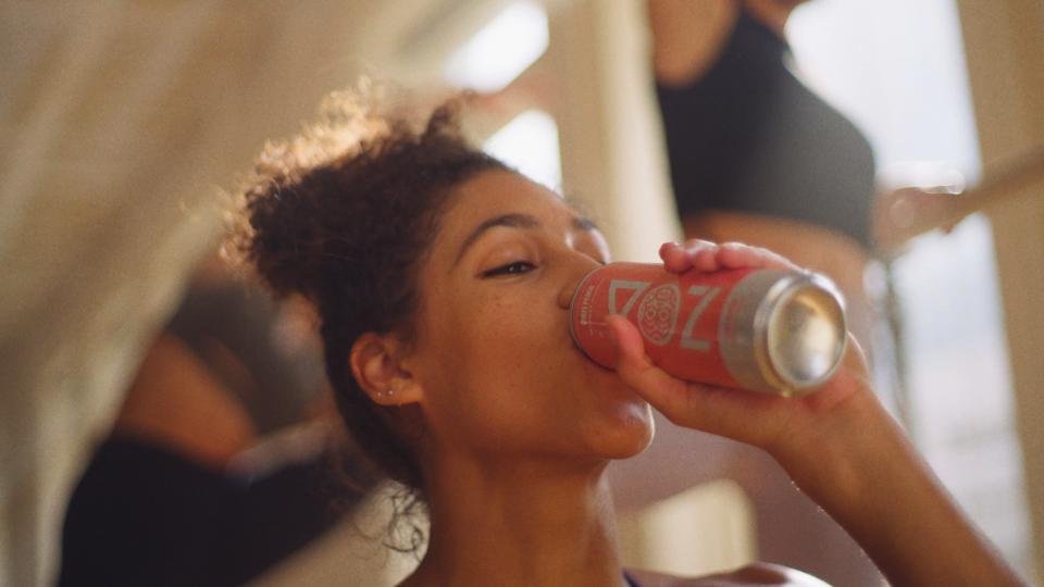 A young woman drinks a can of soda in a casual setting, with another person in the background.