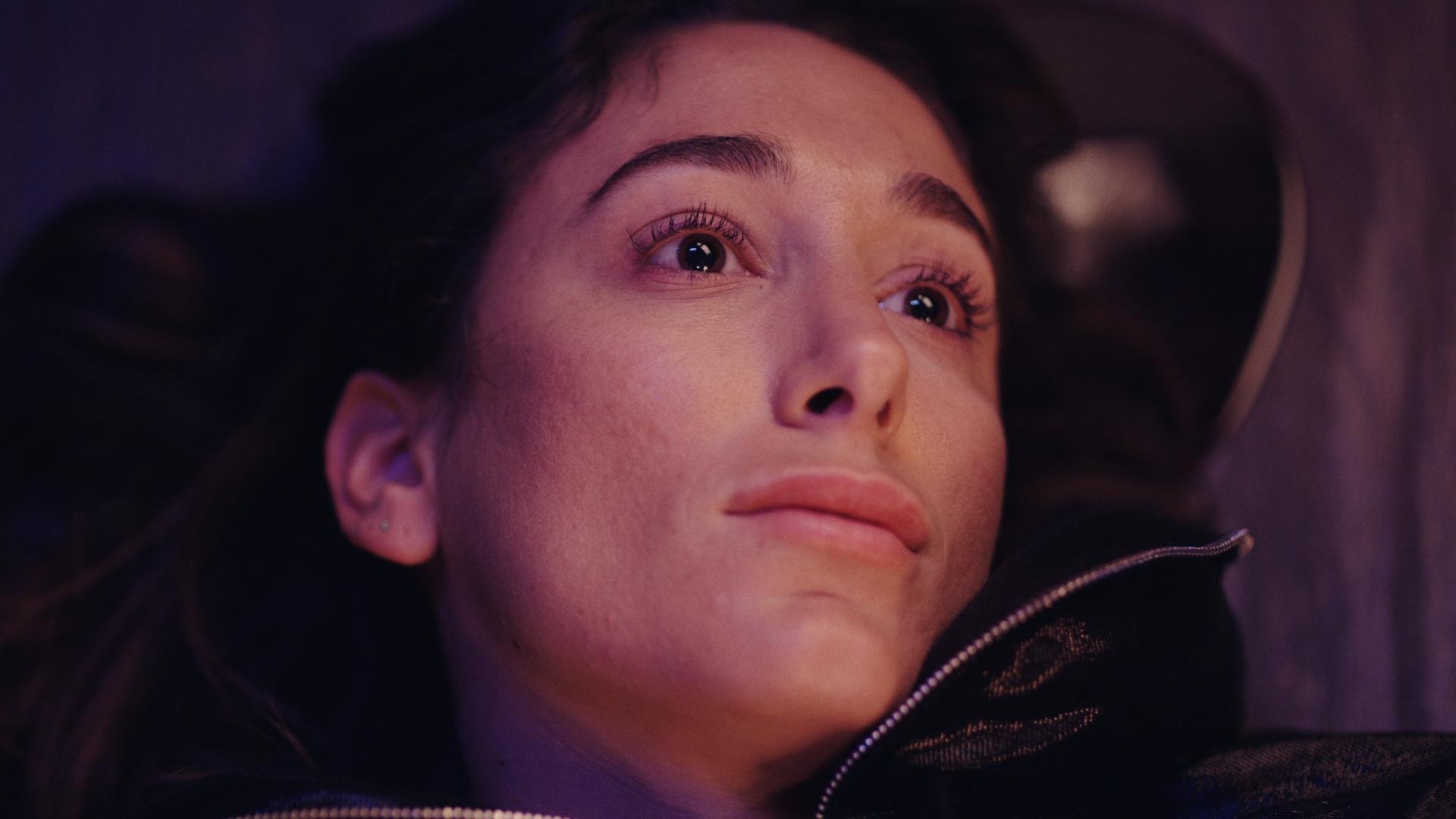 Close-up of a woman lying down with her head resting on a dark surface, looking slightly upwards with a calm expression, illuminated by soft, pinkish-purple lighting.