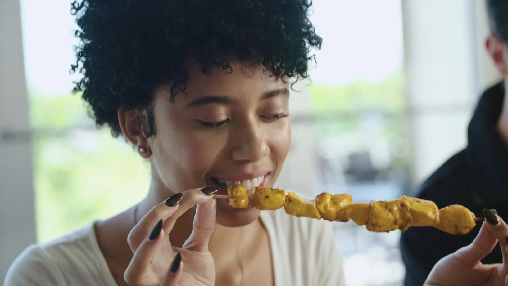 Woman with short curly hair enjoying a skewer of grilled chicken.