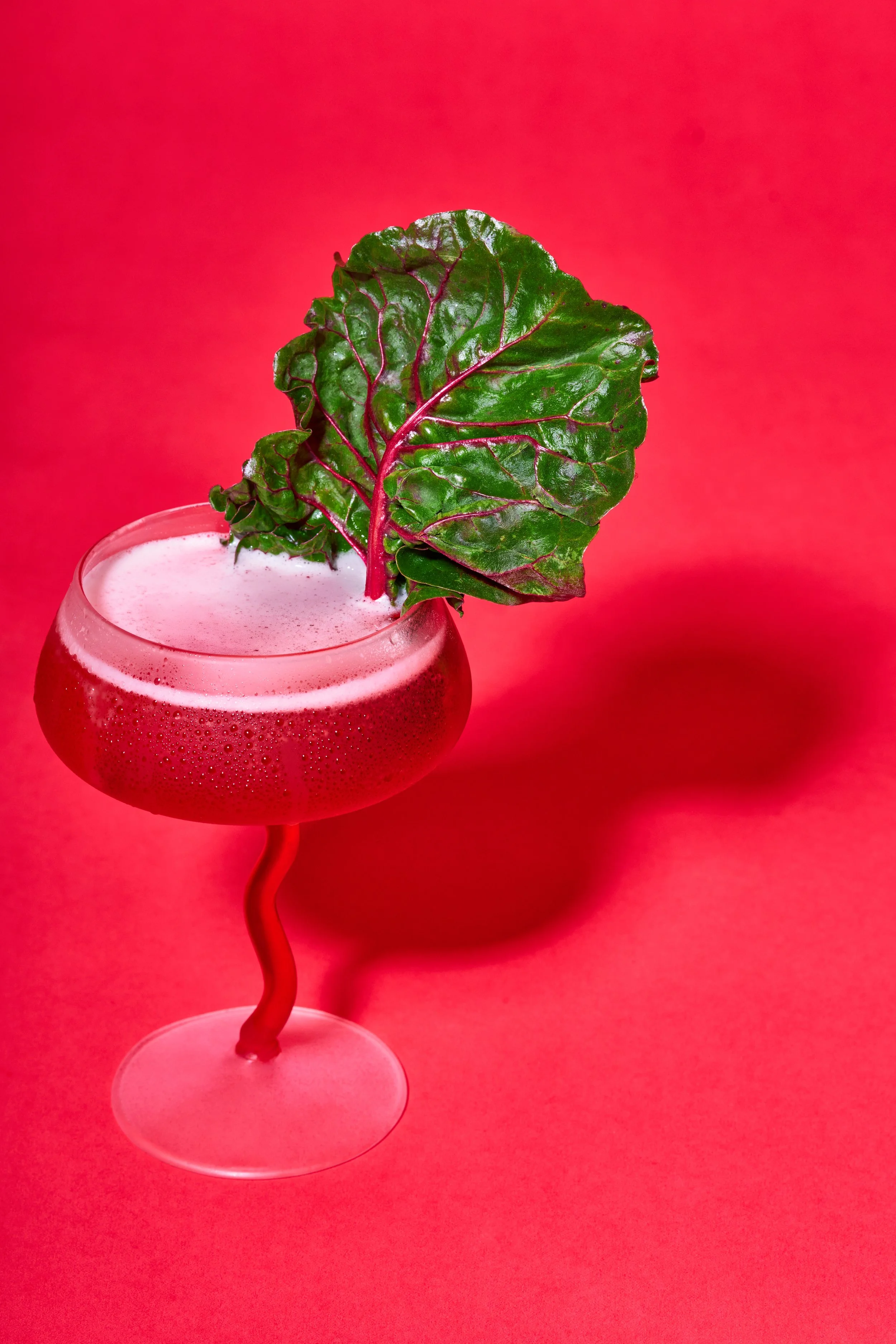 Close-up of a red cocktail in a stemmed glass with a large Swiss chard leaf garnish, set against a red background.