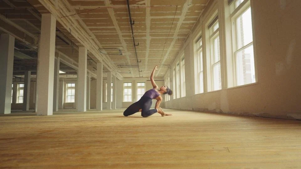 A woman practicing yoga in an unfinished, spacious room with large windows.