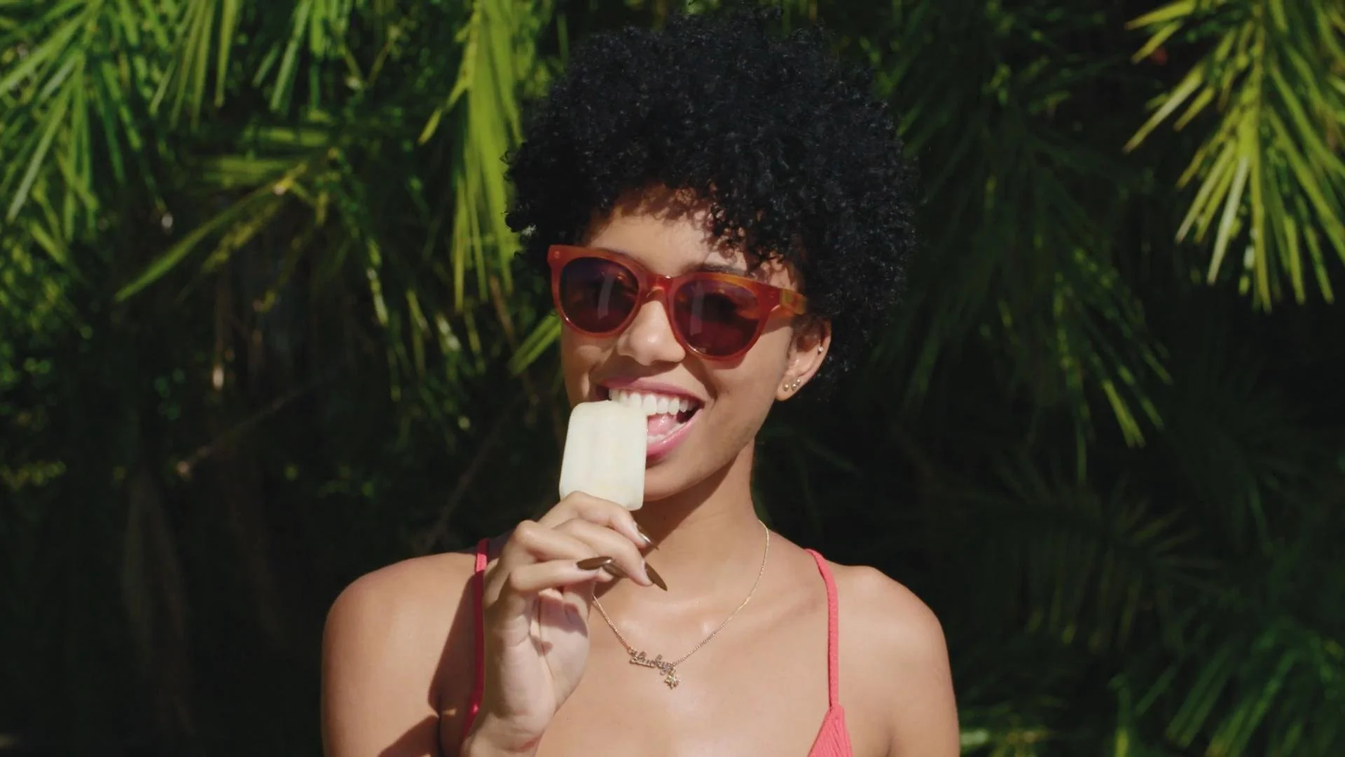 A woman with curly black hair wearing sunglasses and a pink spaghetti strap top, smiling and holding an ice cream bar on a stick, outdoors with green foliage in the background.
