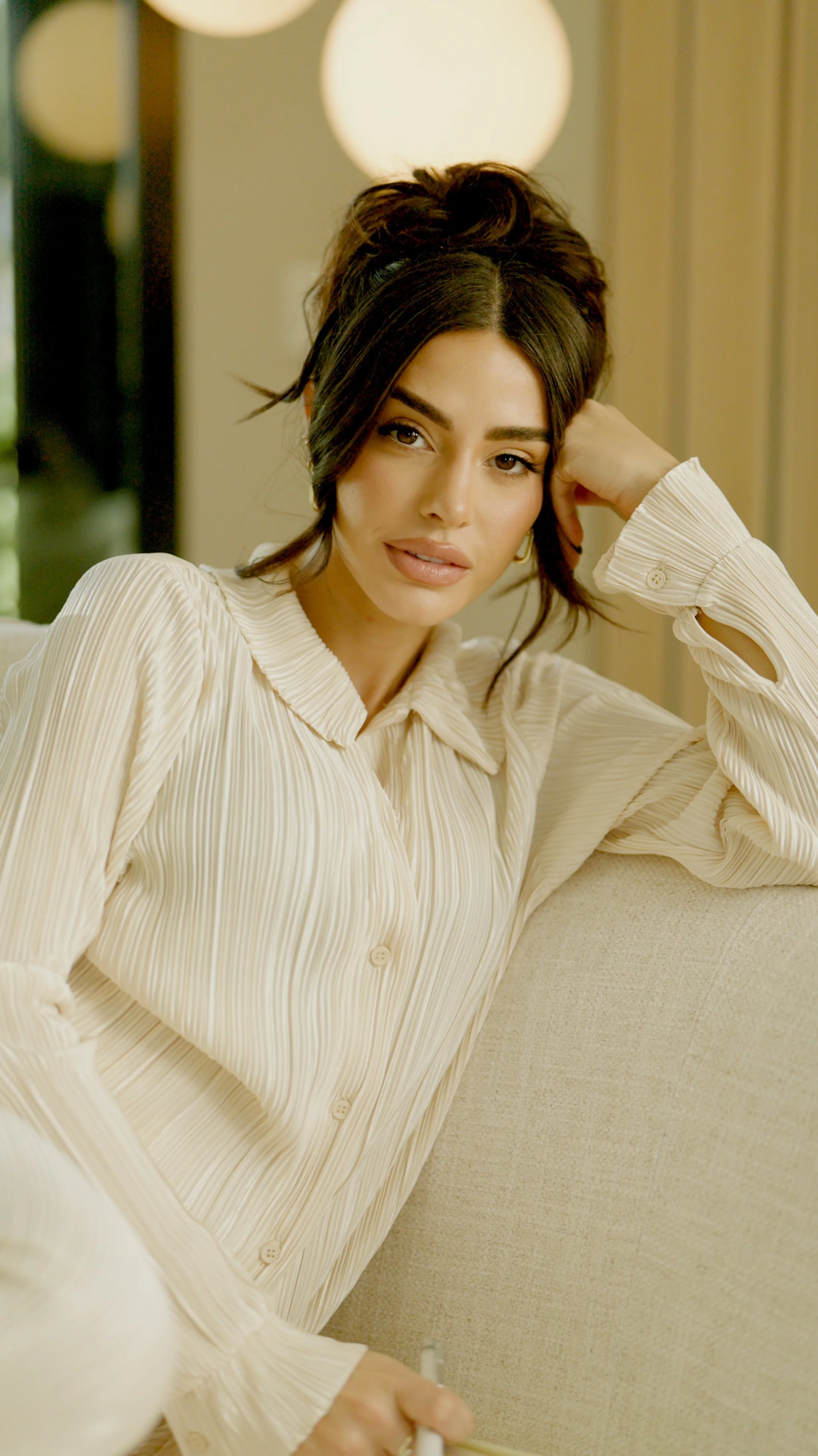 A woman with dark hair styled in an updo, wearing a beige pleated blouse, sitting on a beige sofa and leaning against the armrest, with a relaxed expression and her hand resting behind her head.
