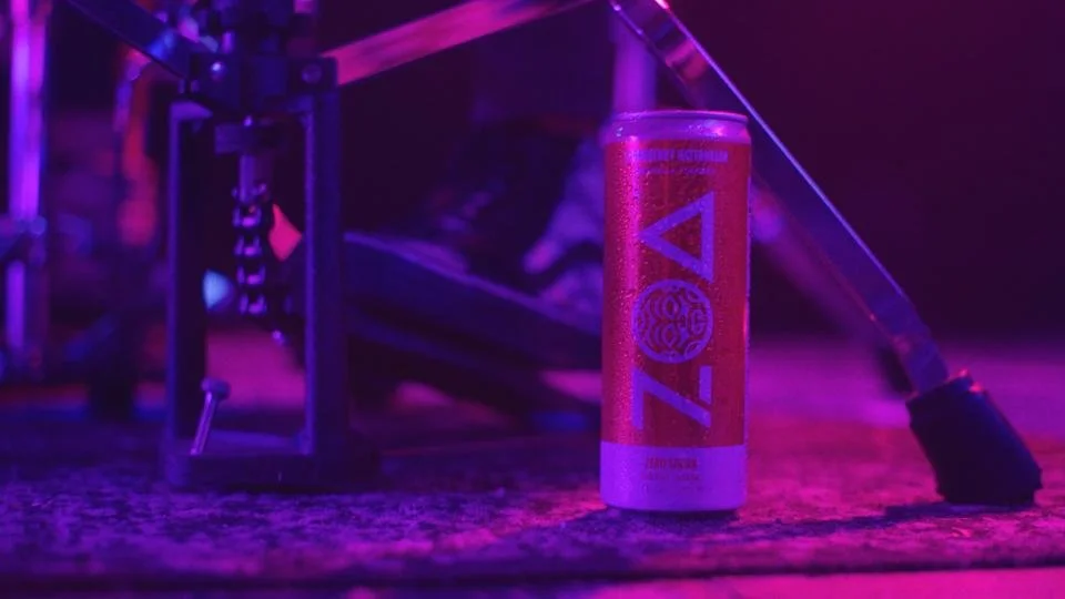 A ZOA energy drink can is placed on a textured surface next to a skateboard with neon purple and pink lighting.