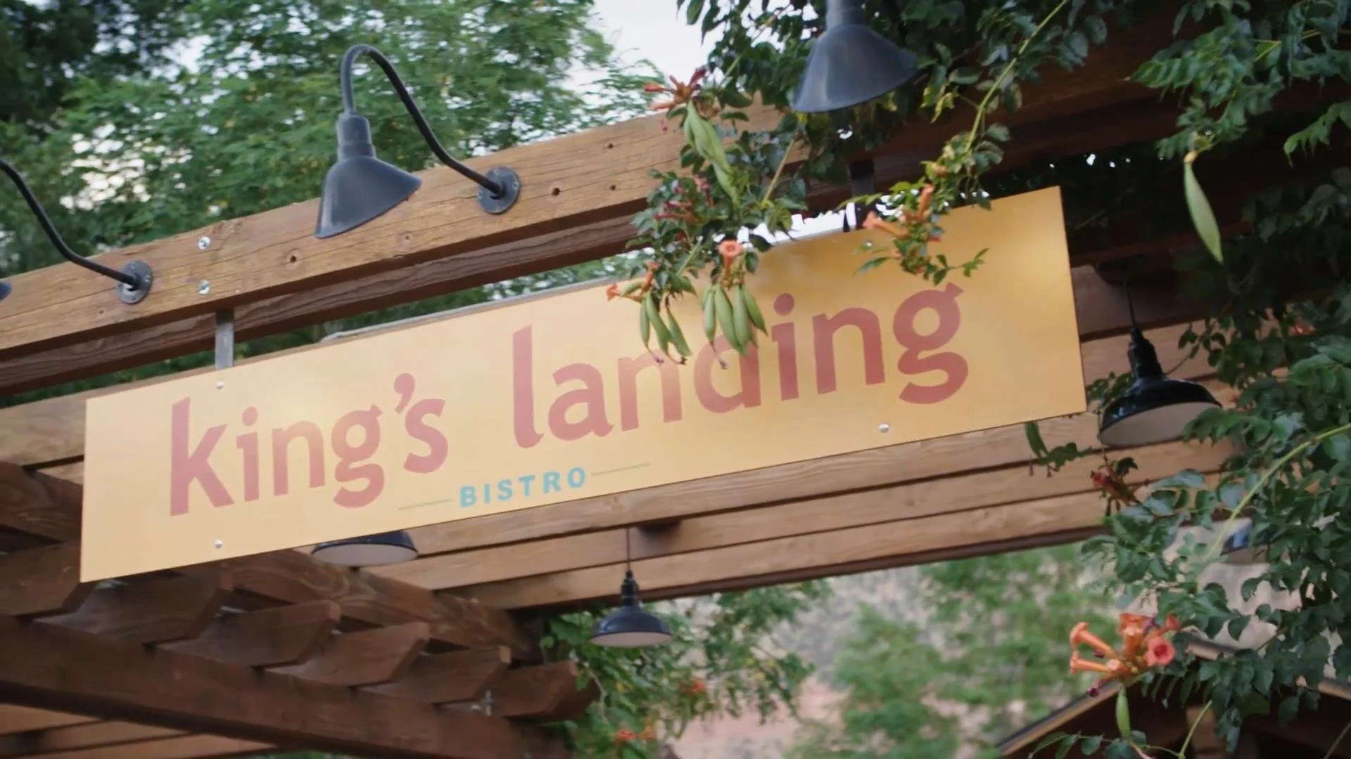 Sign for King's Landing Bistro hanging under a wooden pergola with string lights and surrounded by green foliage and orange flowers.
