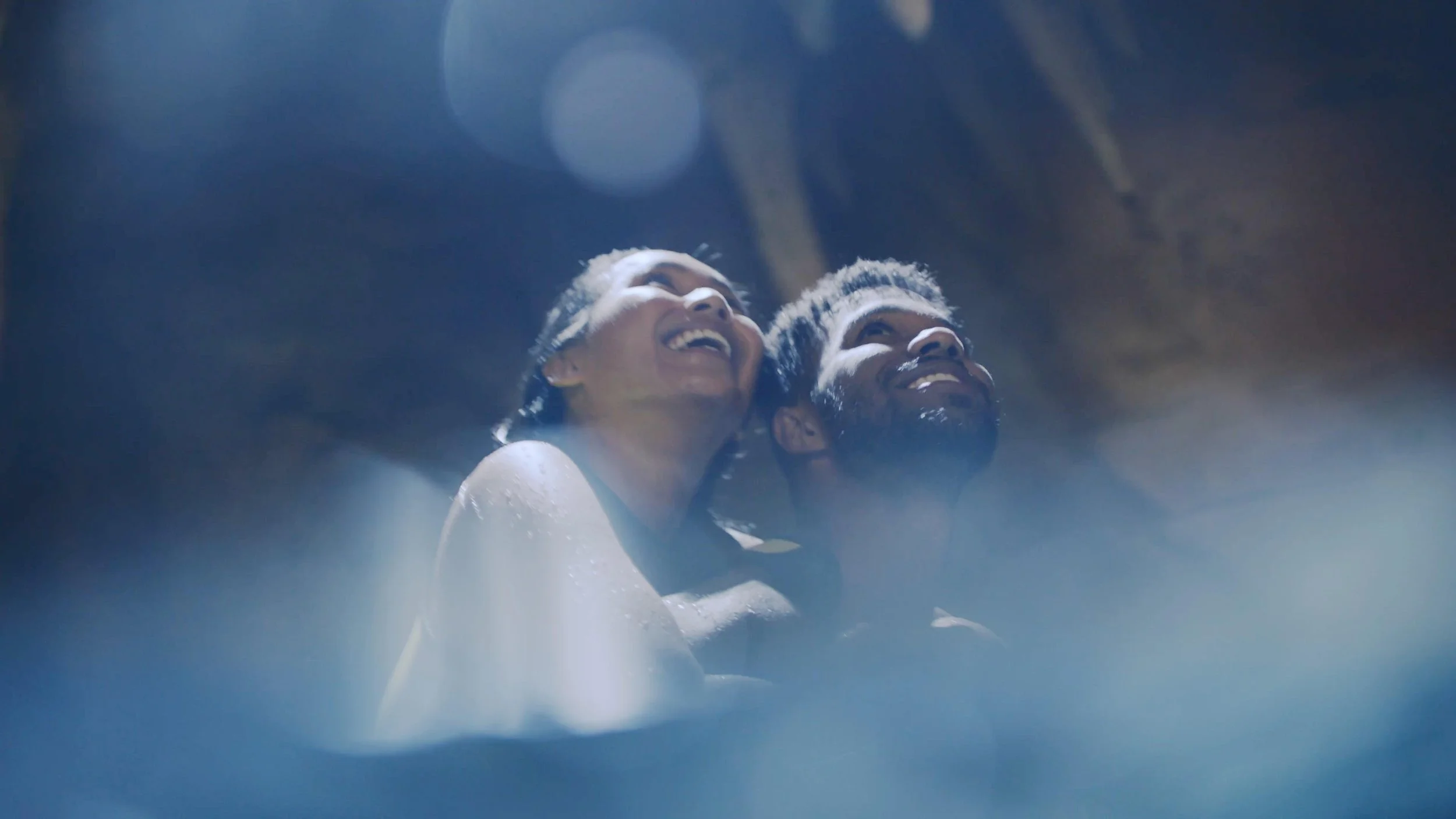 Two people laughing and embracing, with their faces close together, under blue lighting.
