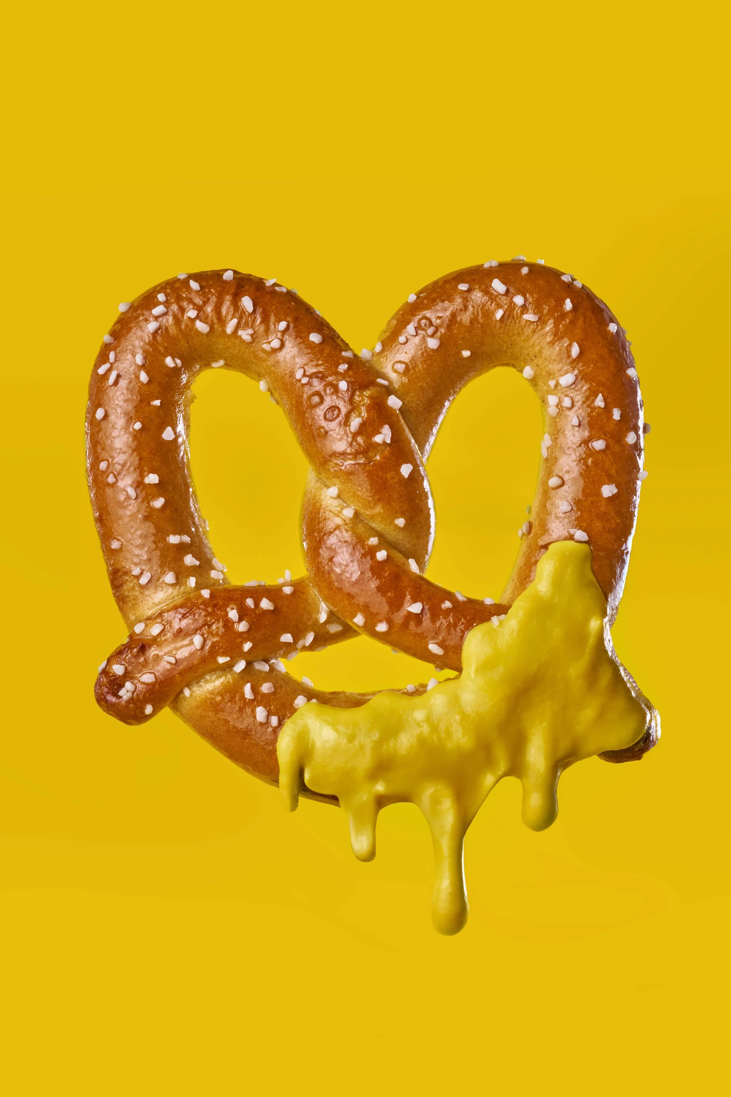 A giant pretzel with coarse salt crystals, dipped in melted cheese, set against a solid bright yellow background.