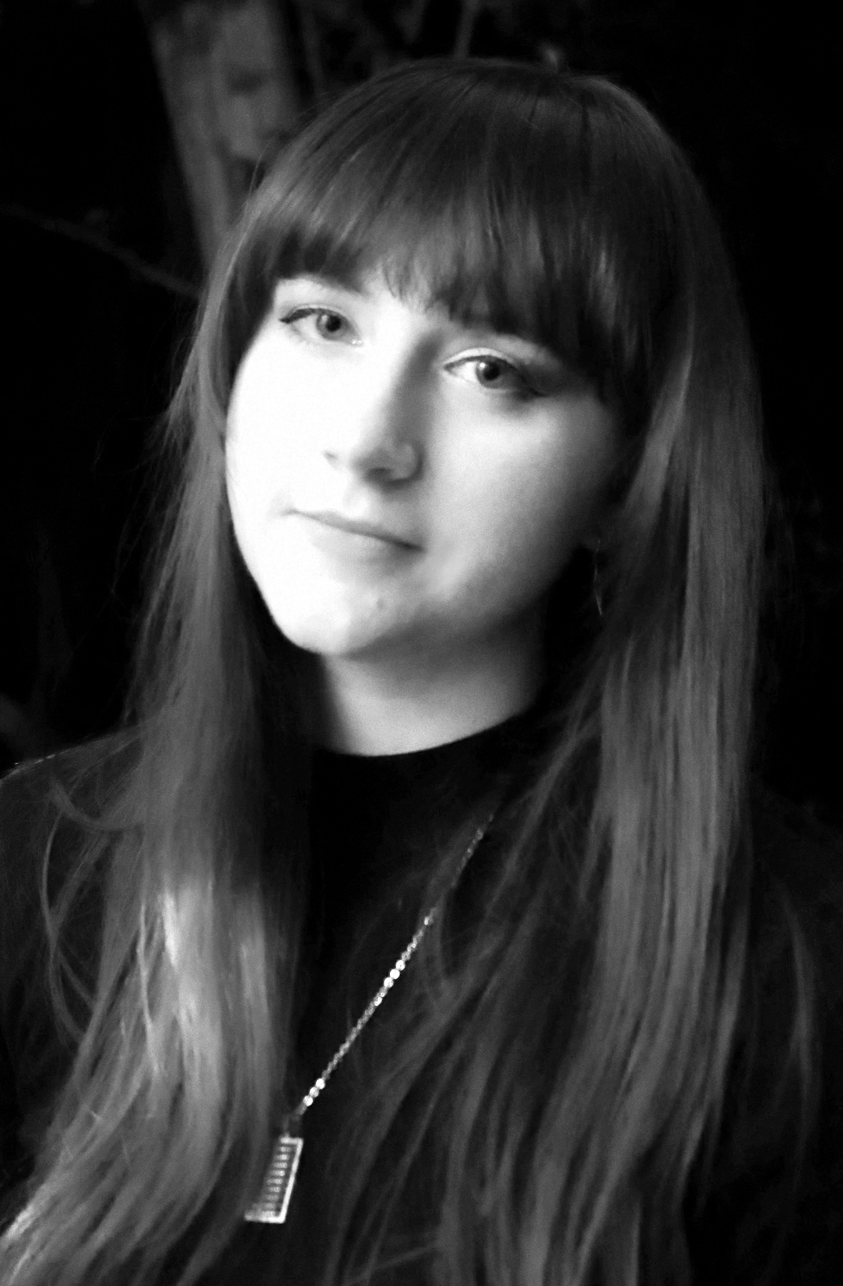 Black and white portrait of a young woman with long hair and bangs, wearing a zip-up jacket with a necklace, looking slightly to the side.