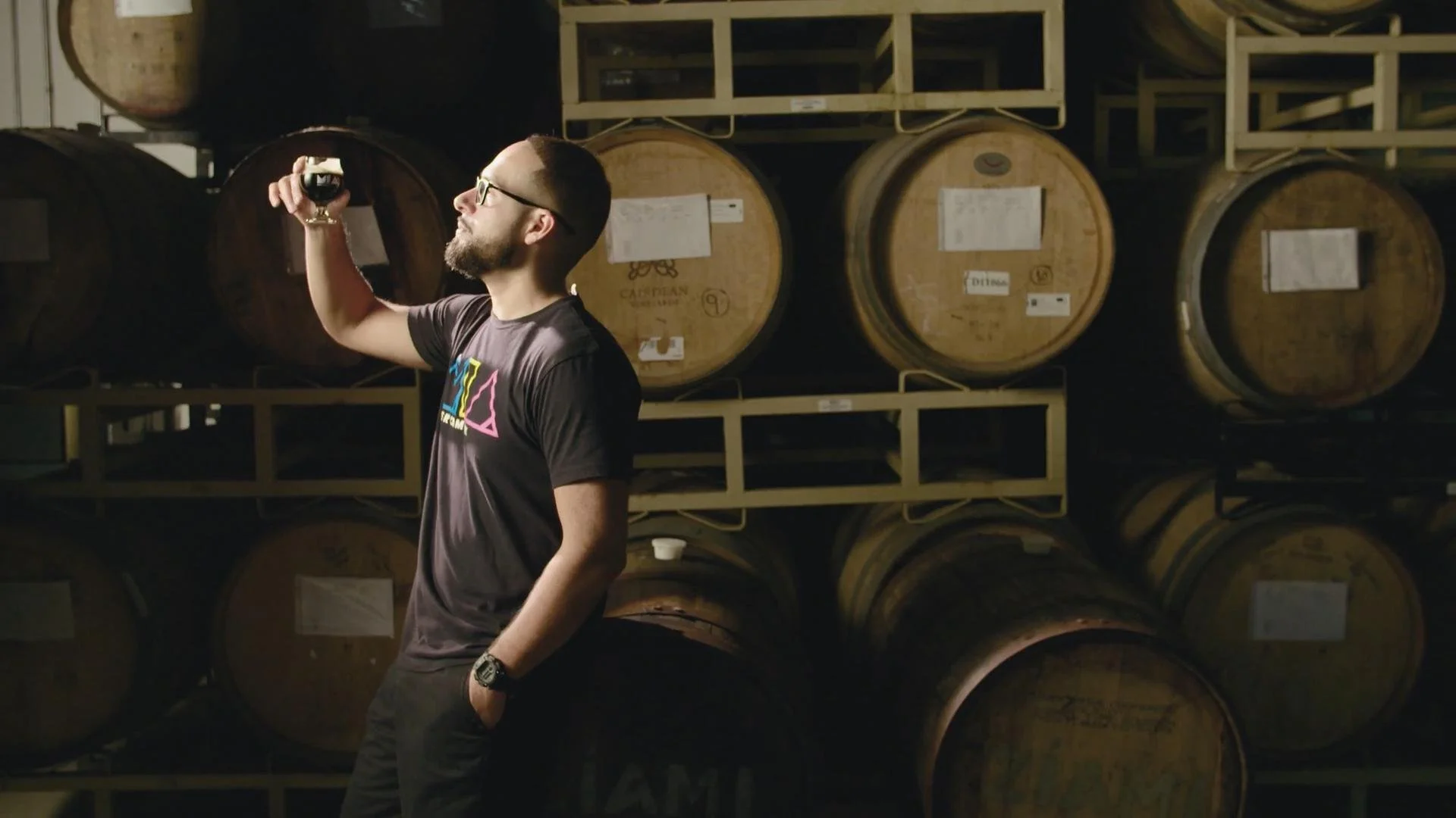 A man with glasses and a beard, wearing a dark t-shirt with colorful text, stands in front of large wooden barrels stacked on metal racks, holding a glass of dark beer up to the light.