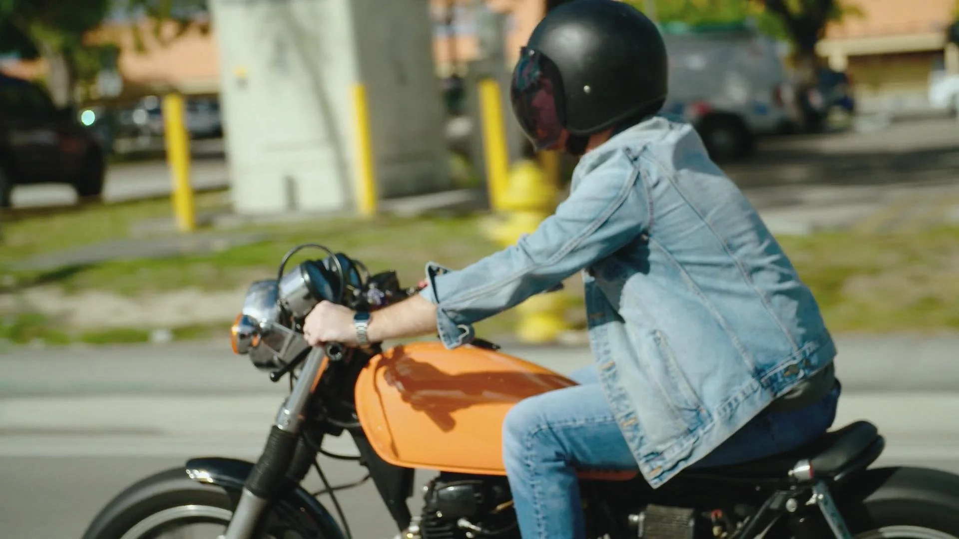 A person wearing a black helmet and a denim jacket riding an orange motorcycle on a city street.