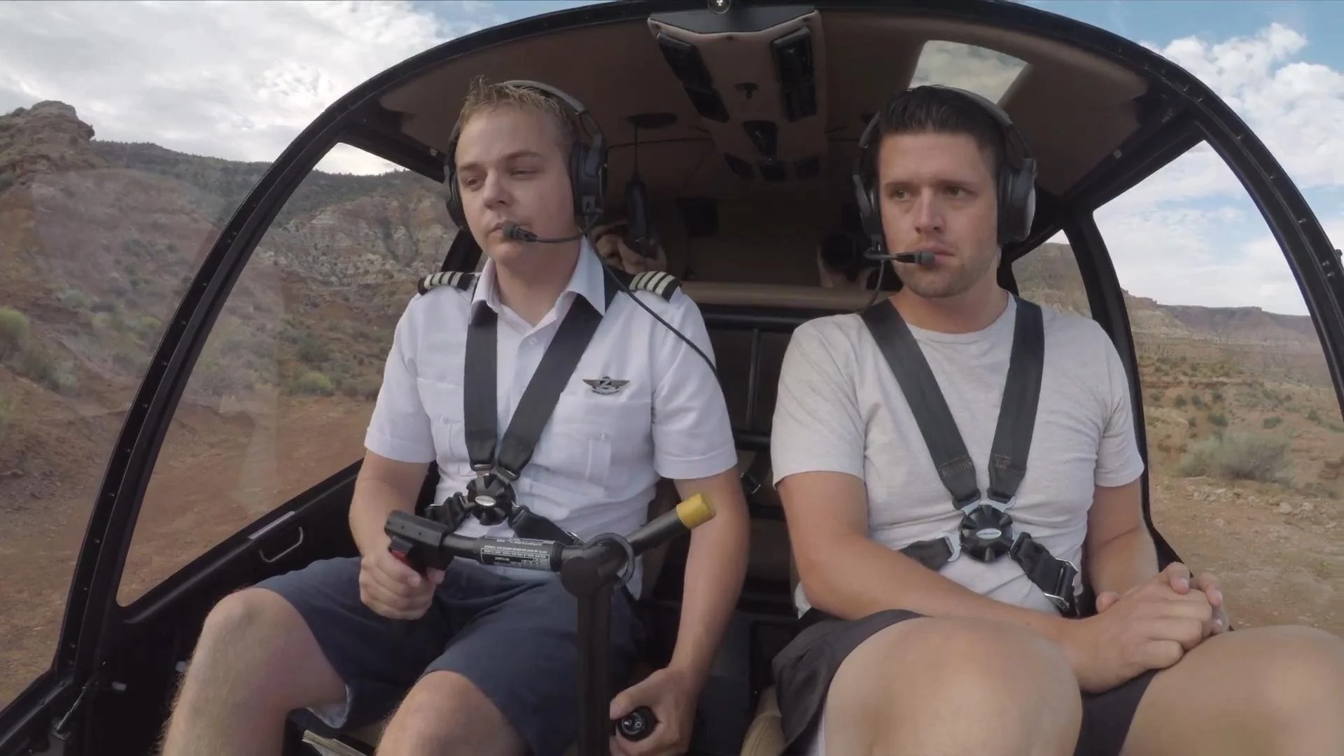 Two men sitting inside a helicopter, one dressed as a pilot with a white uniform and the other in a casual t-shirt, both wearing headsets and harnesses, holding controls, flying over a desert landscape with hills and cloudy sky.