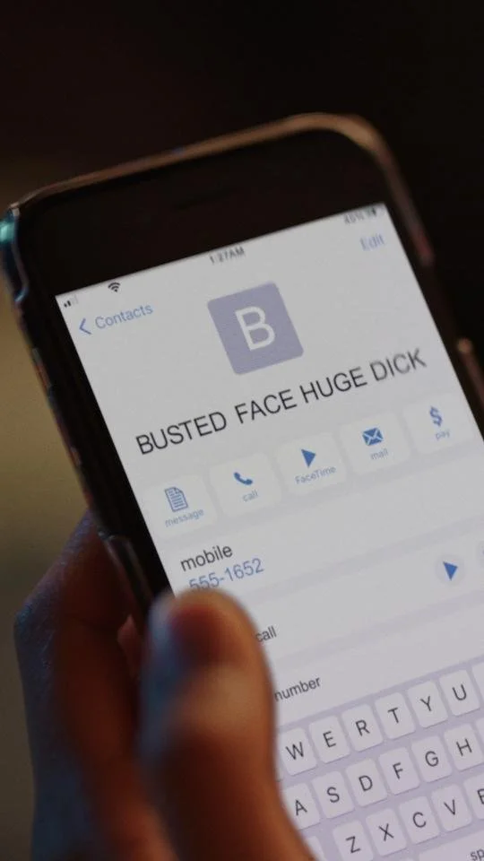Close-up of a smartphone screen displaying a contact named 'BUSTED FACE HUGE DICK' with phone number 555-1652, held in a person's hand.