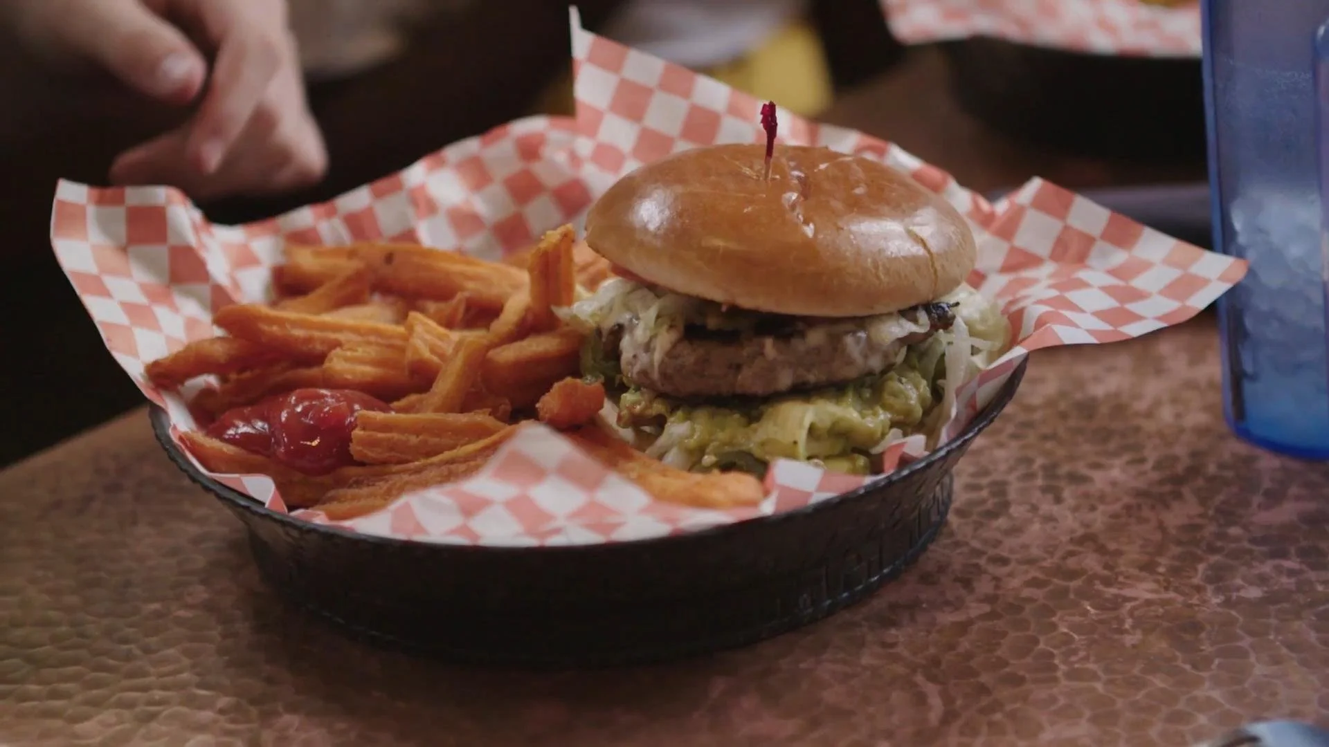 A basket of food with fried sweet potato fries topped with ketchup and a burger with a bun, lettuce, cheese, a sausage patty, and other ingredients, served on red checkered paper.