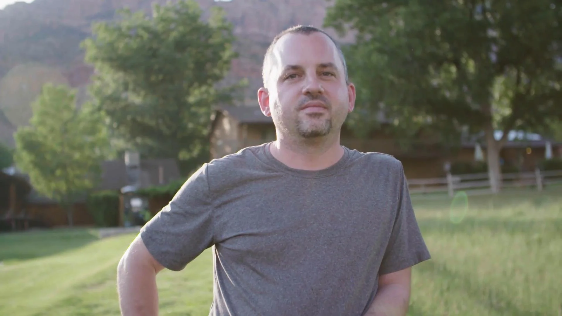 A man with short hair and a goatee, wearing a gray t-shirt, is standing outdoors in a park-like setting with trees and houses in the background during daylight.