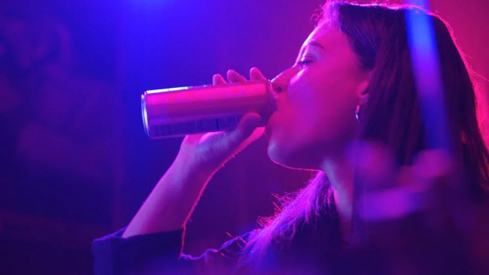 A woman with dark hair drinking from a can in a dimly lit environment with purple and pink lighting.
