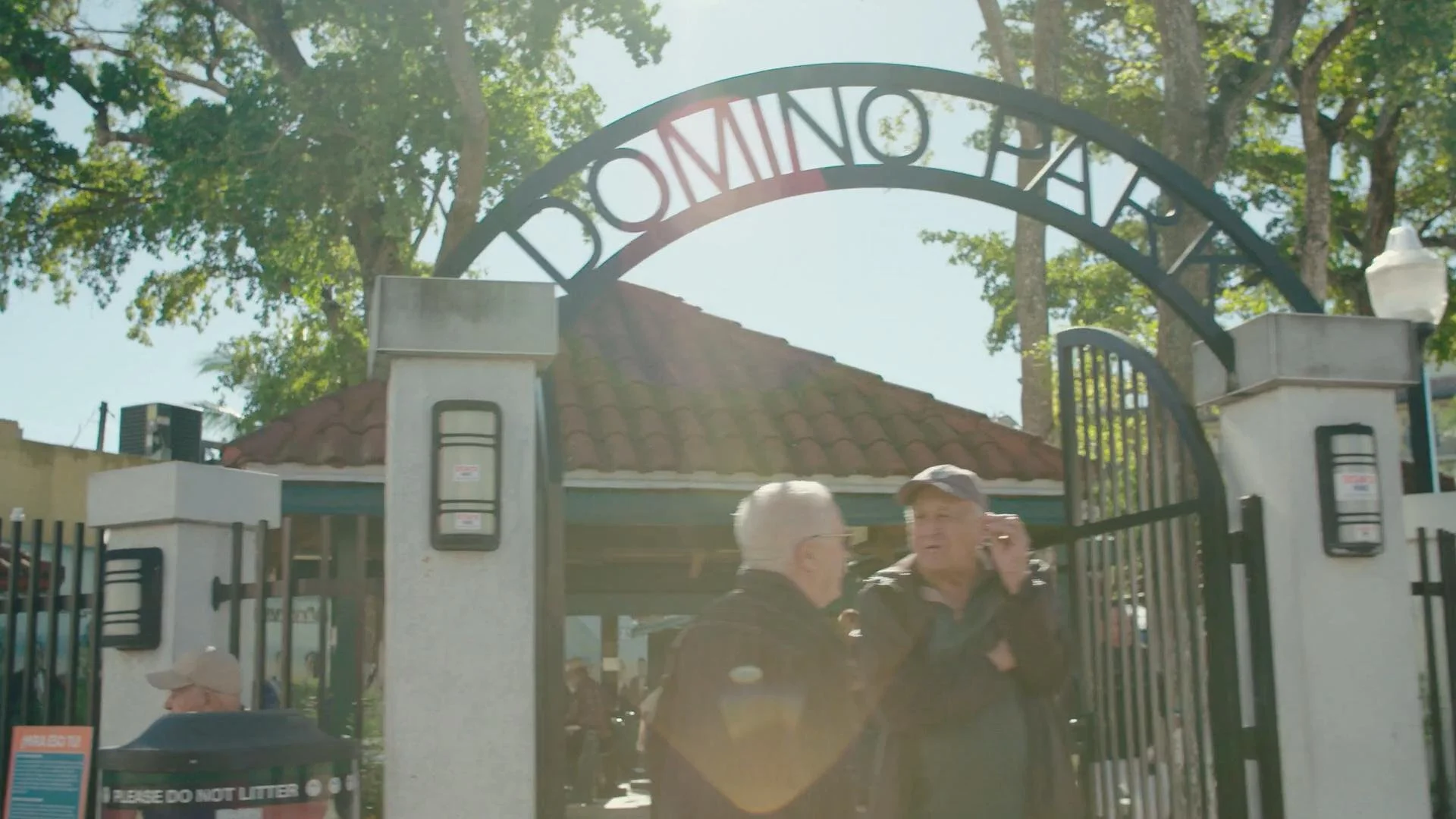 Two men conversing at the entrance of a park or zoo, with a sign reading 'DOMINO PARK' and a gate open behind them, surrounded by trees and sunlight.