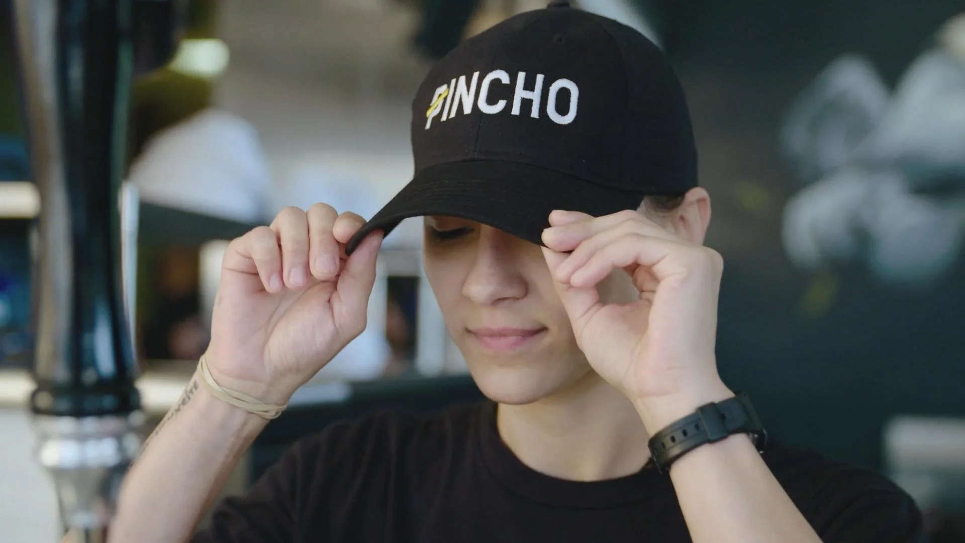 A woman adjusting a black baseball cap with the word 'PINCHO' embroidered on it. She is in a casual setting and wearing a black shirt, a wristwatch, and a wristband.