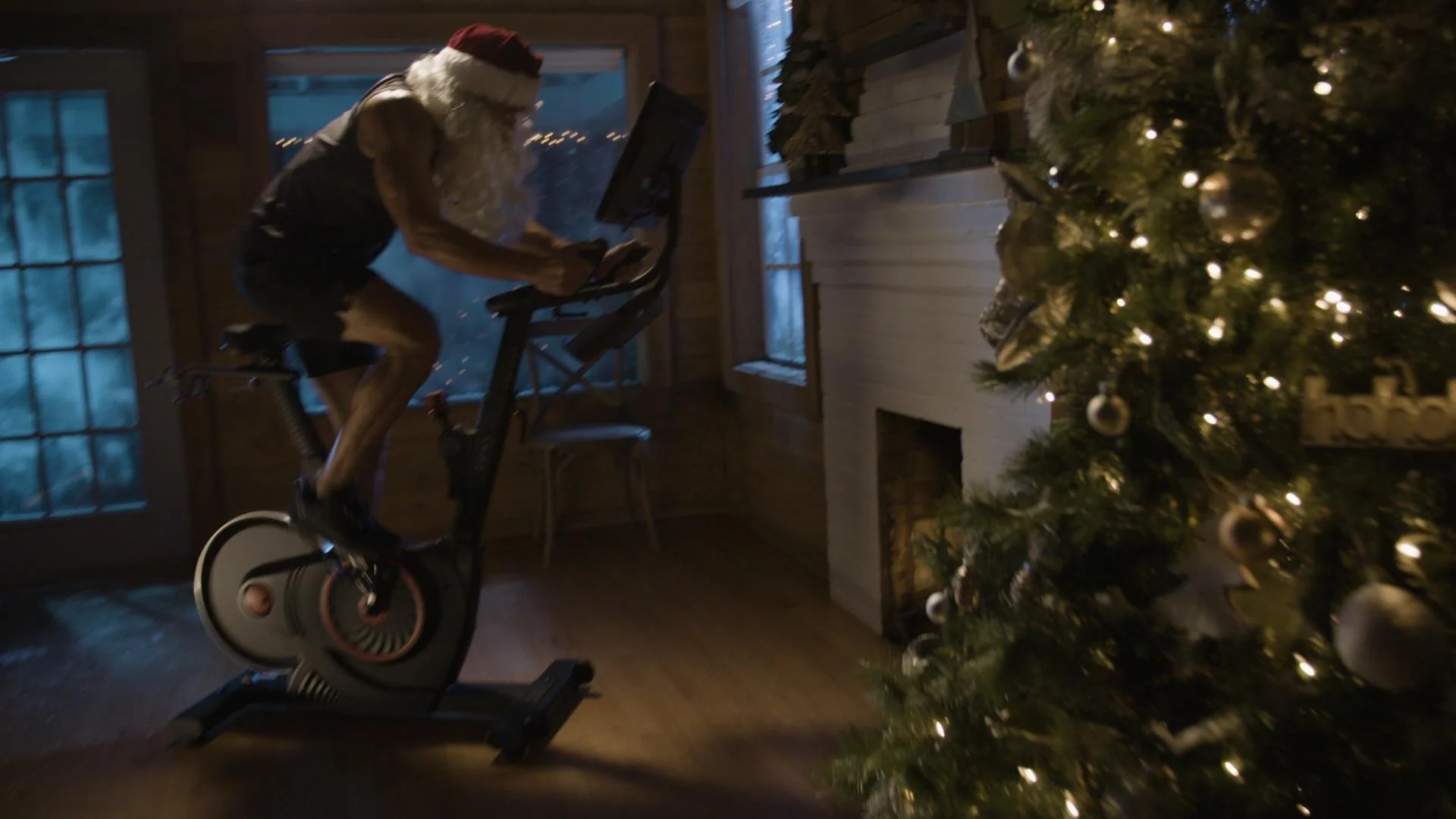 A person dressed as Santa Claus riding a stationary exercise bike inside a house decorated for Christmas, with a decorated Christmas tree in the foreground.